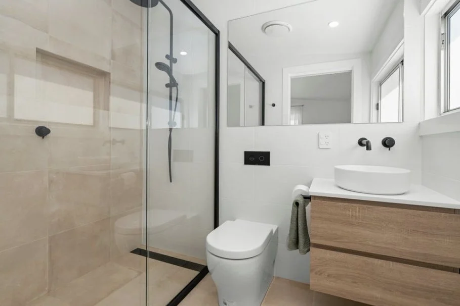 Modern bathroom with glass shower enclosure, wall-mounted toilet, white countertop with round vessel sink, and wooden vanity cabinet. Natural light from a window.