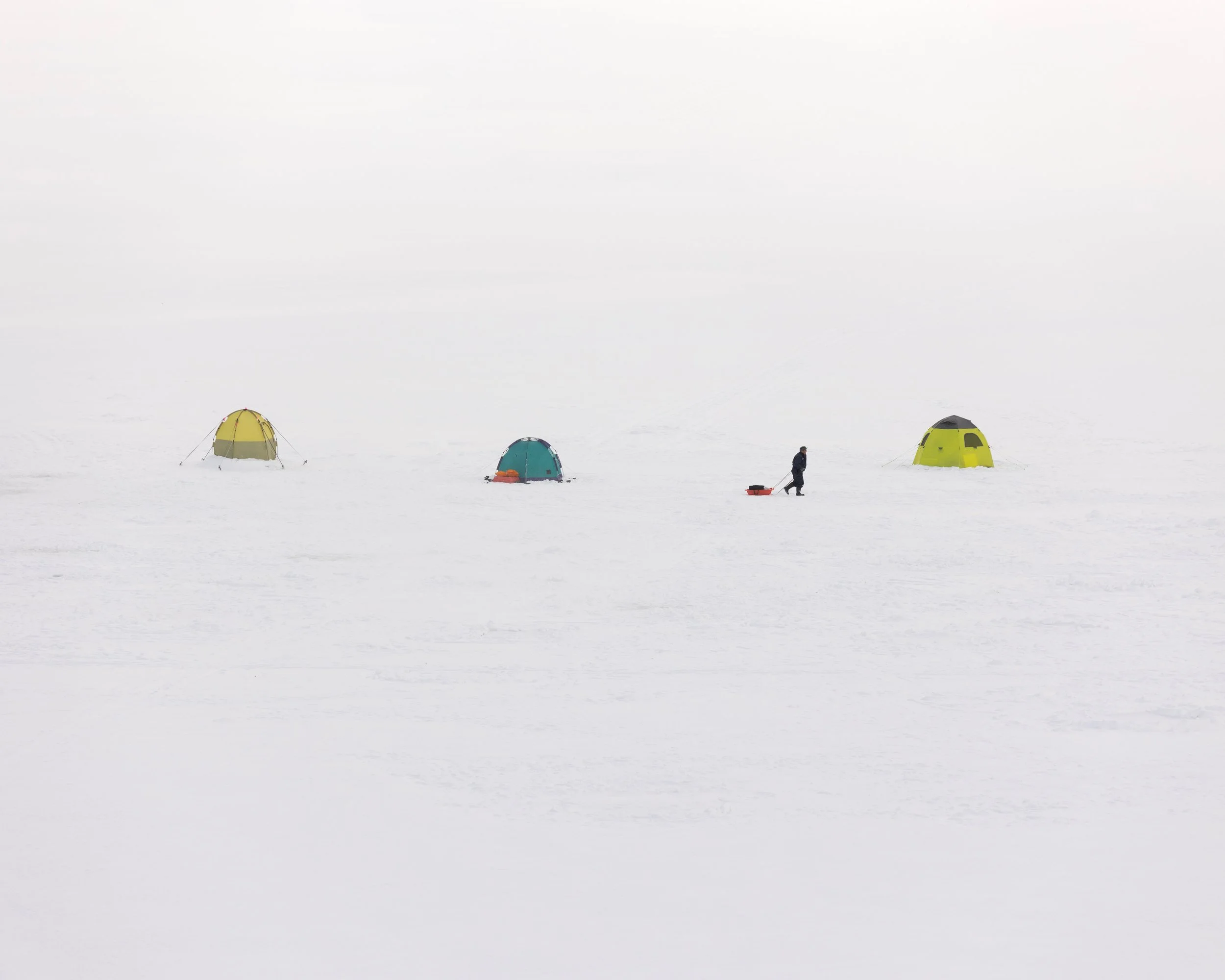 A person pulling a sled on a snowy, open landscape with three yellow and blue tents in the background.