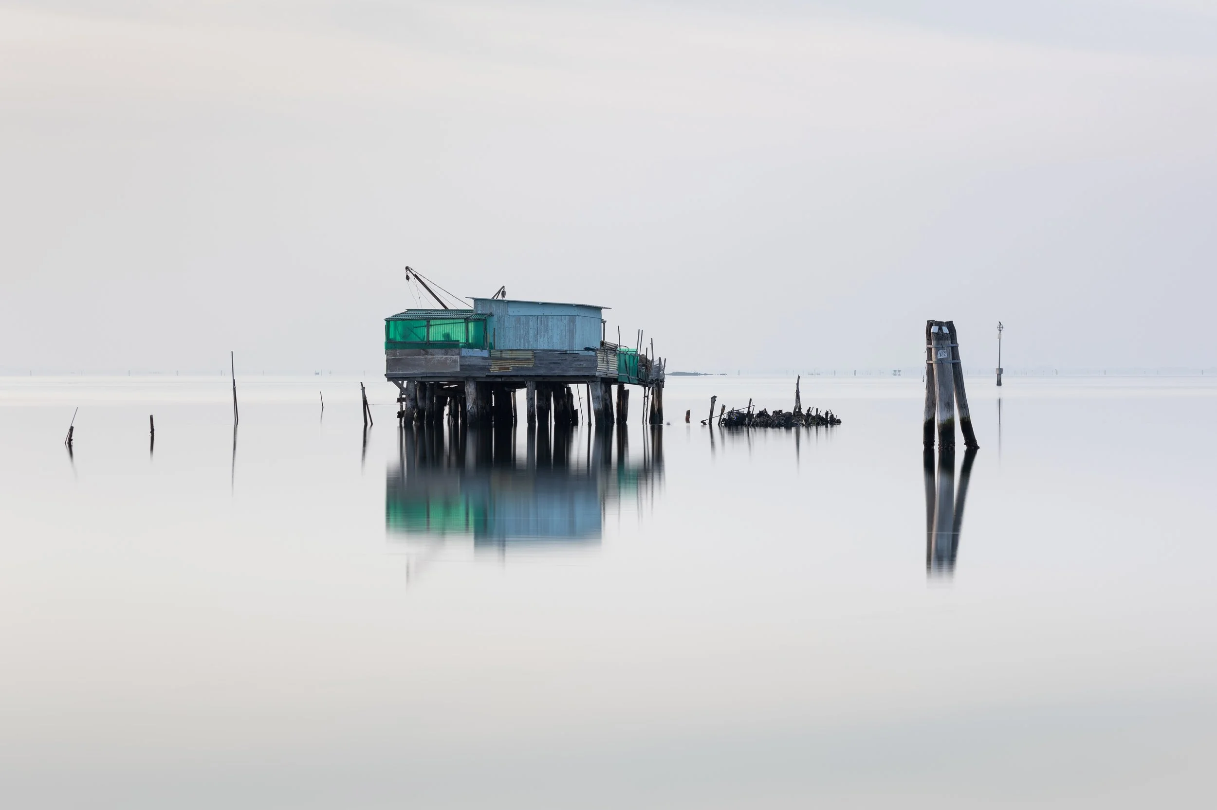 A stilt house over calm water, with wooden posts and remnants of old structures, under a cloudy sky.