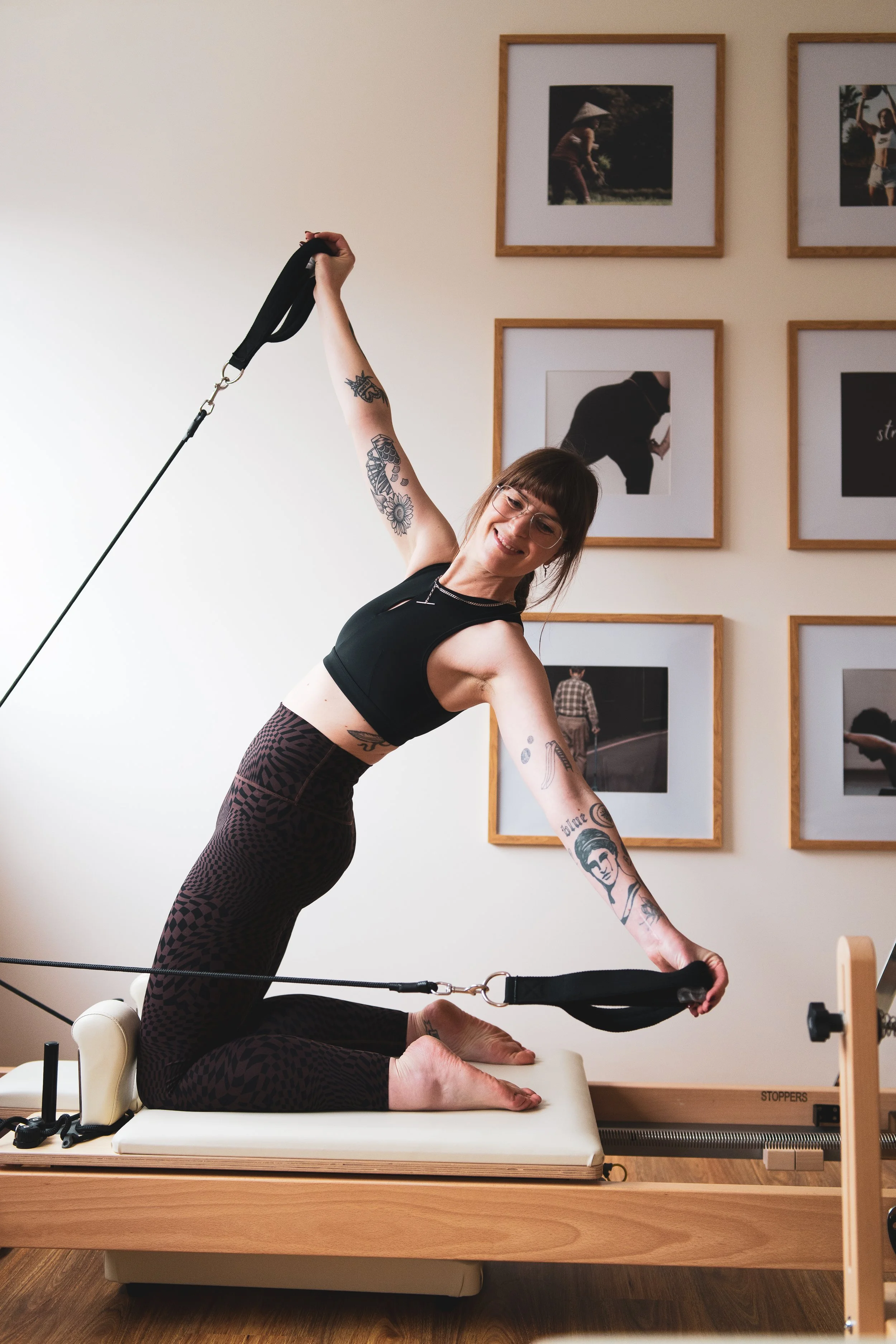 Woman performing Pilates or yoga on a reformer machine, kneeling with straps. She is smiling and wearing glasses, a black sports bra, and leopard print leggings, with framed pictures on the wall behind her.