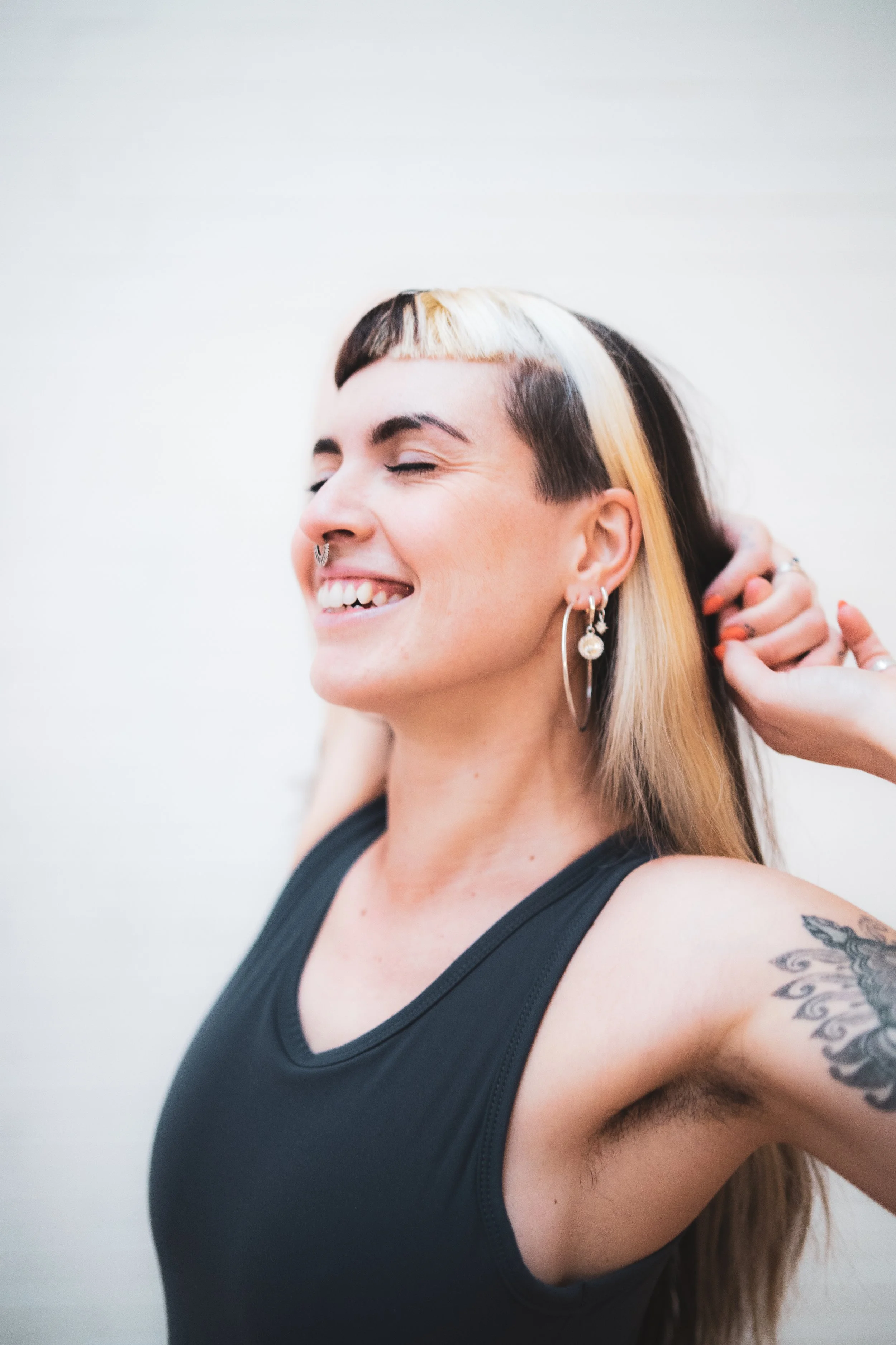 A woman with black and blonde hair, smiling with eyes closed, adjusting her hair, wearing earrings and a black tank top, showing a tattoo of a butterfly on her upper arm.