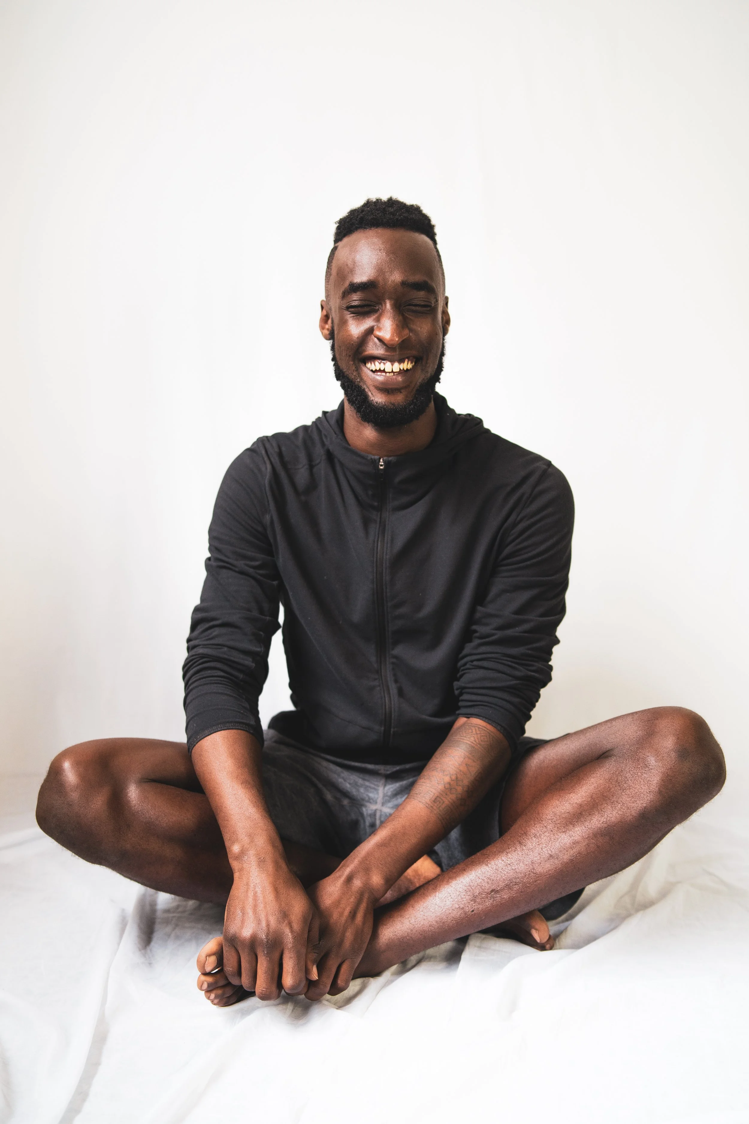 A young man sitting cross-legged on a white sheet, smiling with eyes closed, wearing a black zip-up hoodie and black shorts, against a plain white background.