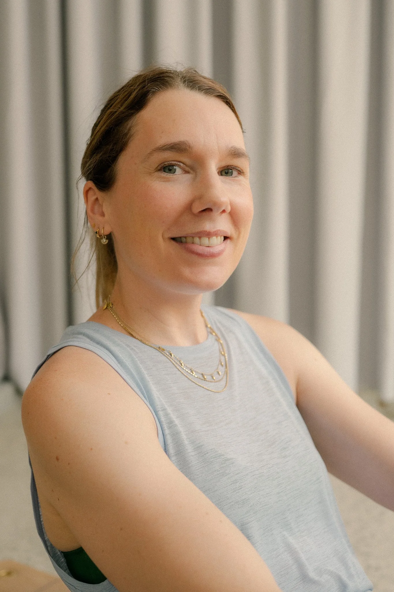 A woman with fair skin and light brown hair, smiling, wearing a sleeveless gray top and layered gold necklaces, with gray curtains in the background.