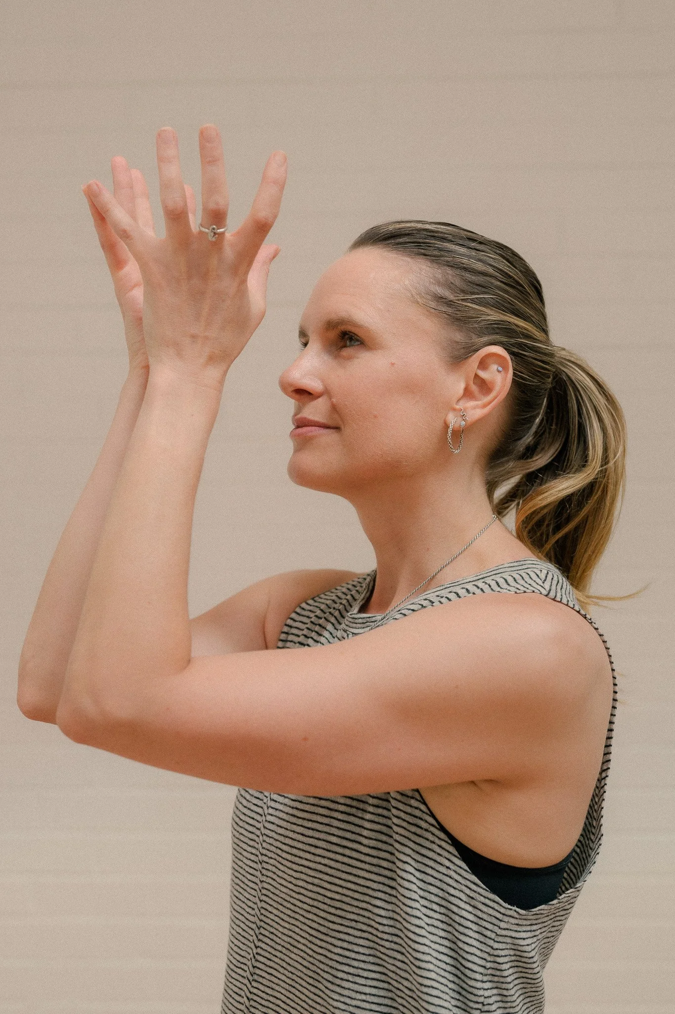Woman with blonde hair tied back, wearing hoop earrings and a sleeveless striped top, reaches up with both hands, gazing slightly upward.