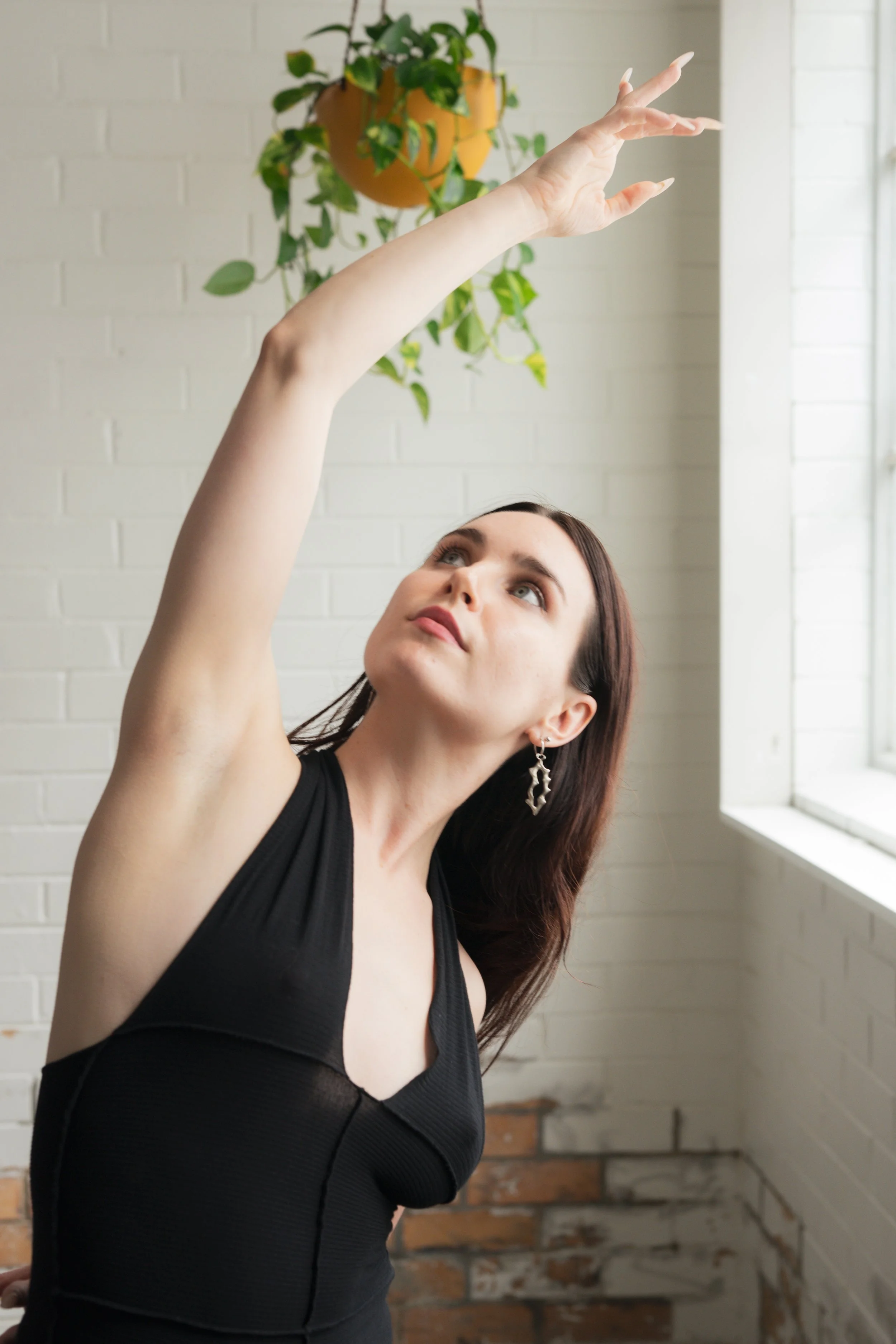 A woman with dark brown hair wearing a black sleeveless top and silver earrings, reaching upward with her right arm near a window with white brick wall background and a potted plant hanging from the ceiling.