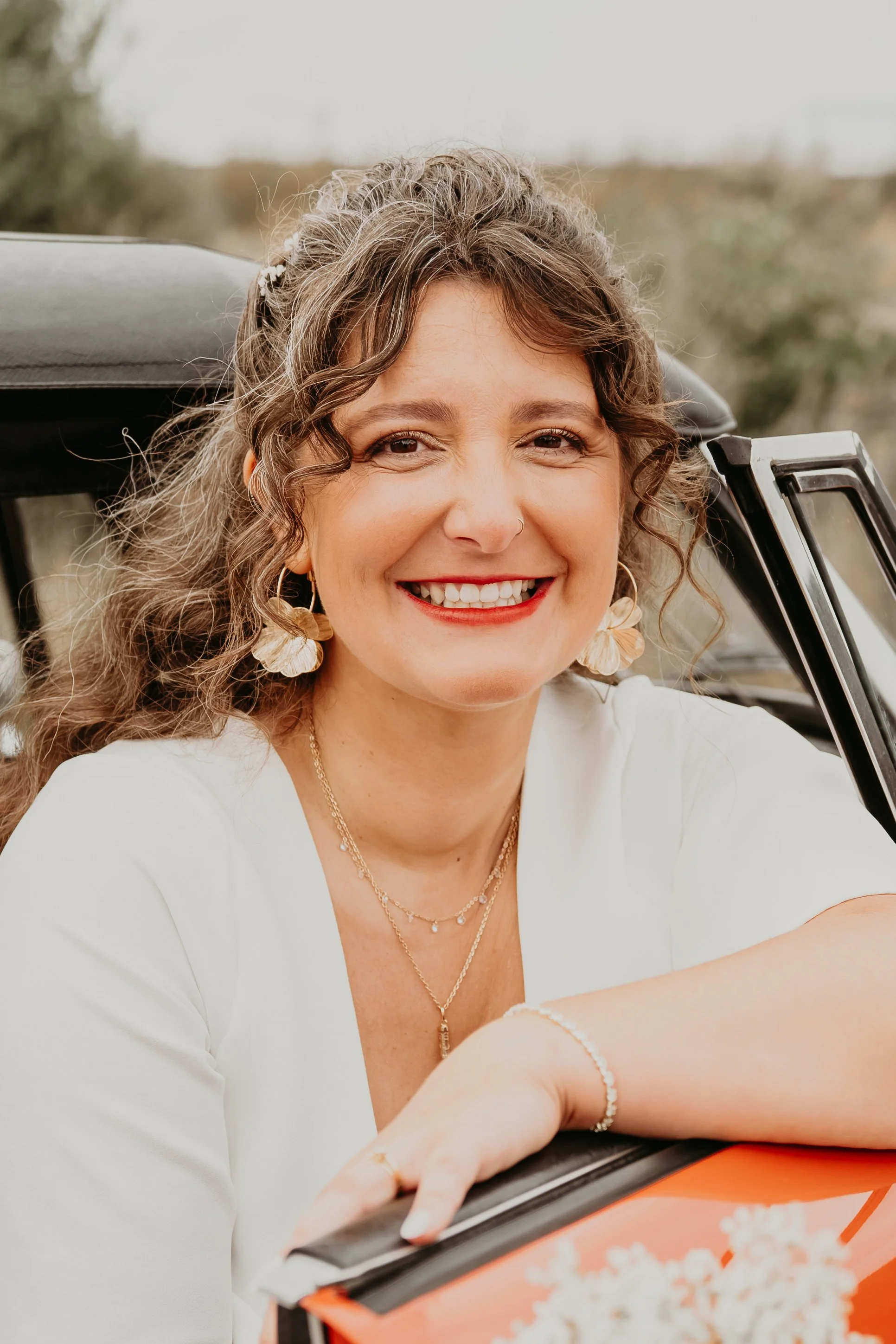 Femme souriante de face avec des boucles d'oreilles en forme de feuilles, portant un collier, un bracelet et un maquillage naturel, posant dans une voiture.