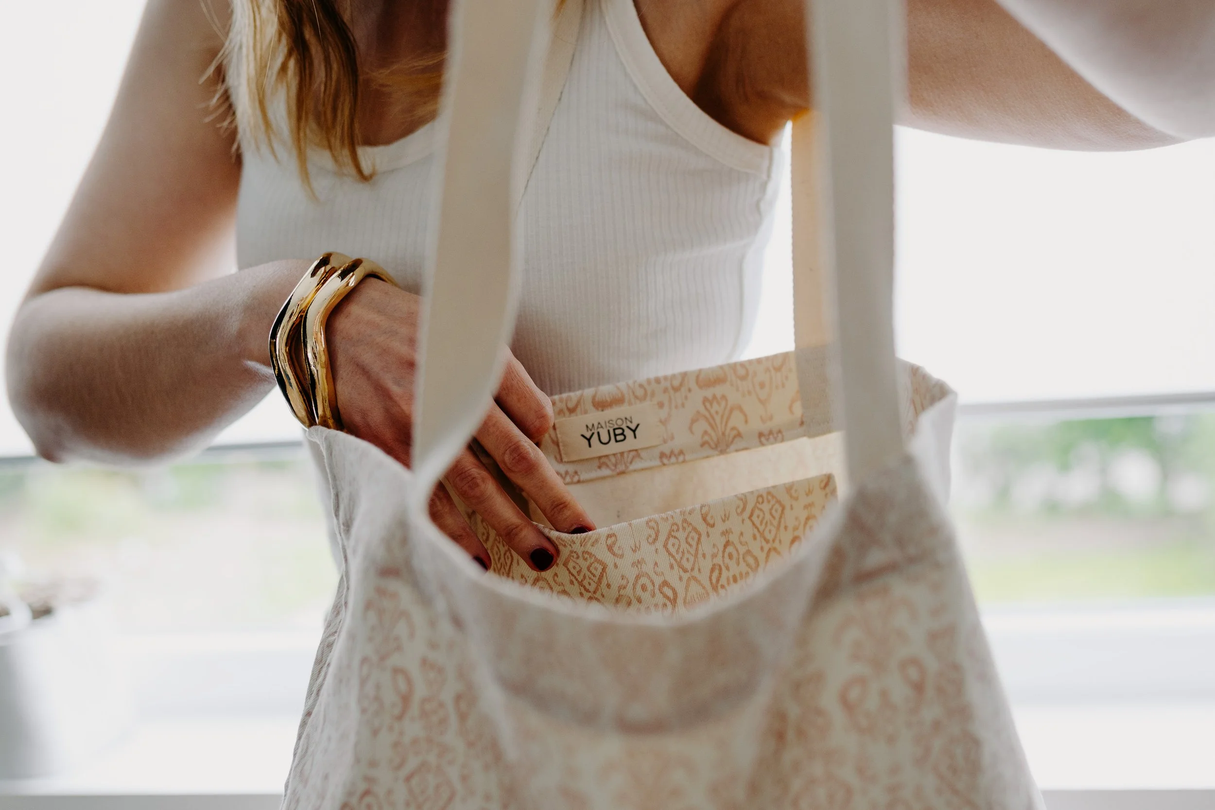 Une femme met la main dans un sac à main en tissu beige avec un motif. Elle porte un bracelet en or et un haut blanc côtelé, debout près d'une fenêtre avec vue sur un espace vert extérieur.