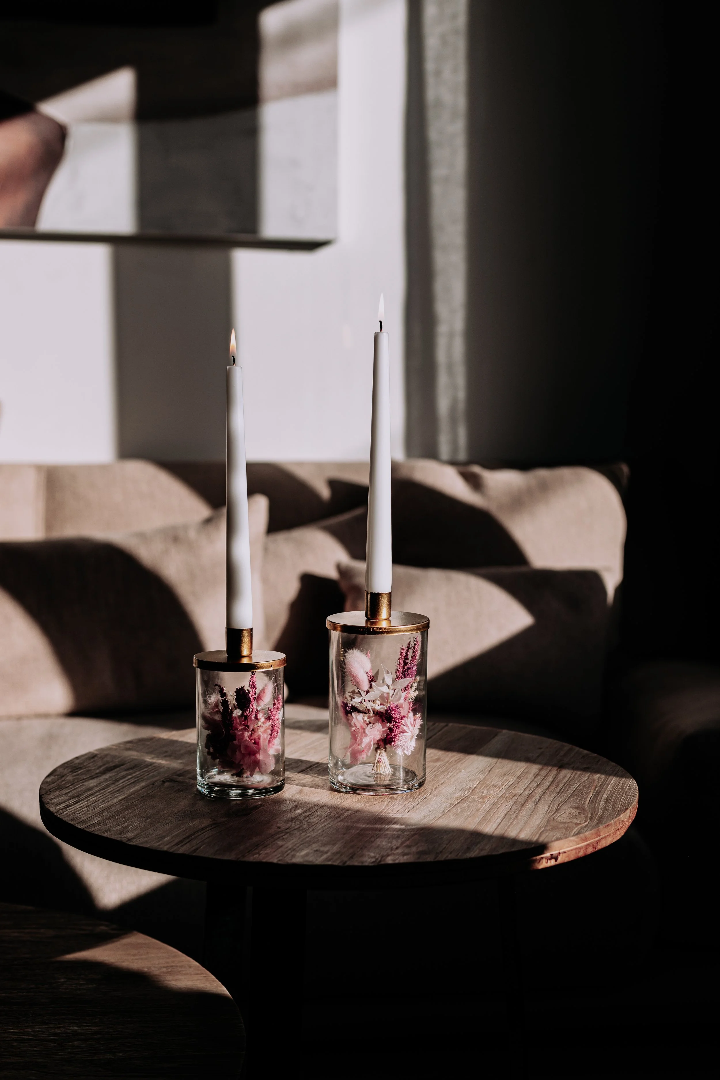Deux bougeoirs en verre avec fleurs roses et rouges, chacun avec une bougie blanche allumée, placés sur une table en bois dans une pièce faiblement éclairée.