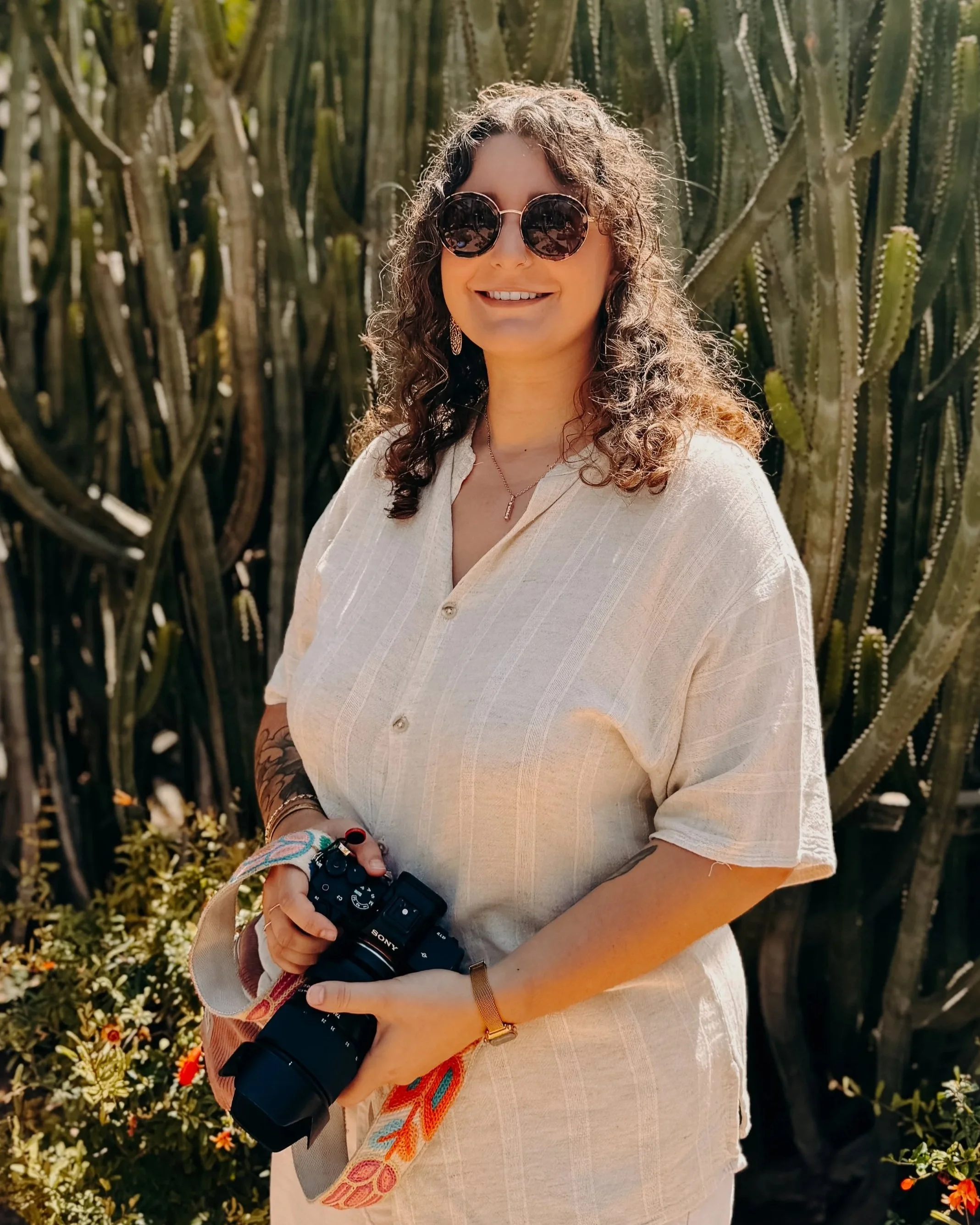 Jeune femme souriante portant des lunettes de soleil rondes, une chemise beige, tenant un appareil photo, avec des cactus en arrière-plan.