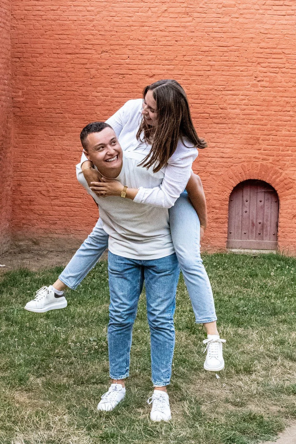 Deux personnes, un homme et une femme, jouent de manière joyeuse dans un jardin avec un mur rouge en arrière-plan. L'homme porte un t-shirt blanc et un jean, la femme porte une chemise blanche et un jean clair. La femme est assise sur le dos de l'homme, qui la supporte, tous deux sourient et semblent s'amuser.