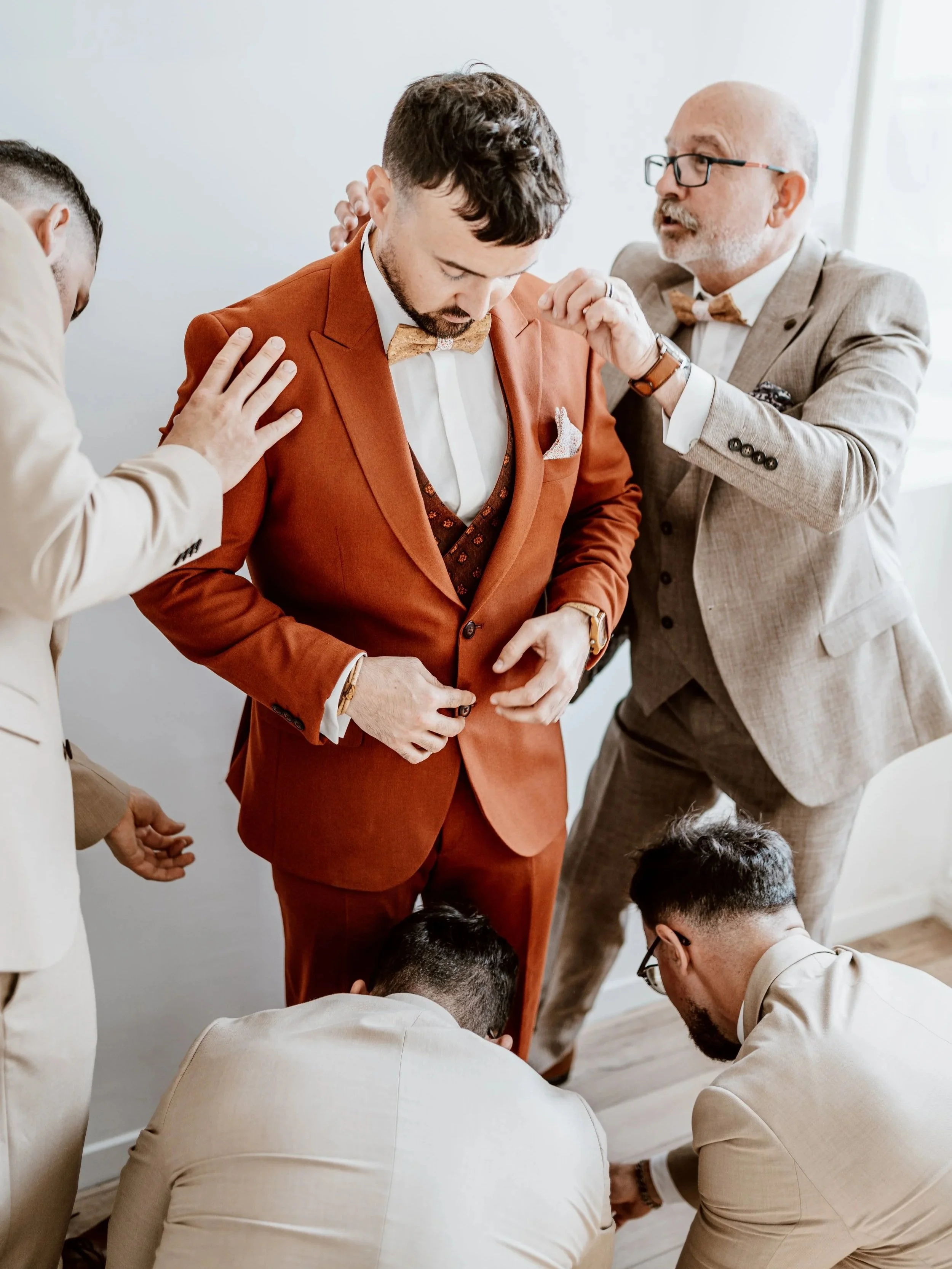 Un groupe de cinq hommes, dont un en costume orange, en train de se préparer pour un événement formel, probablement un mariage, avec des conseils donnés par d'autres.