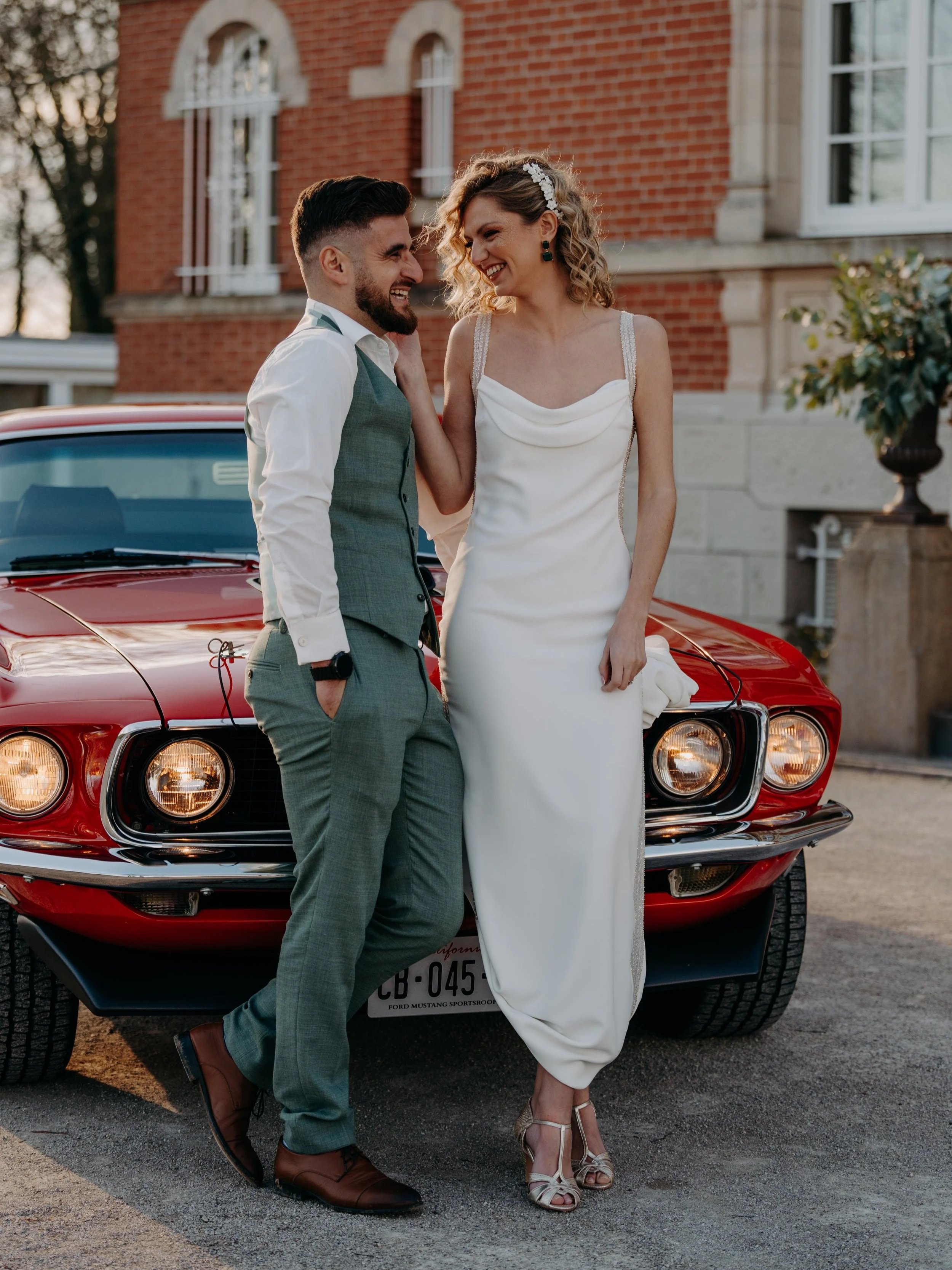 Un couple en tenue de mariage posant devant une voiture rouge, avec une maison en brique en arrière-plan.