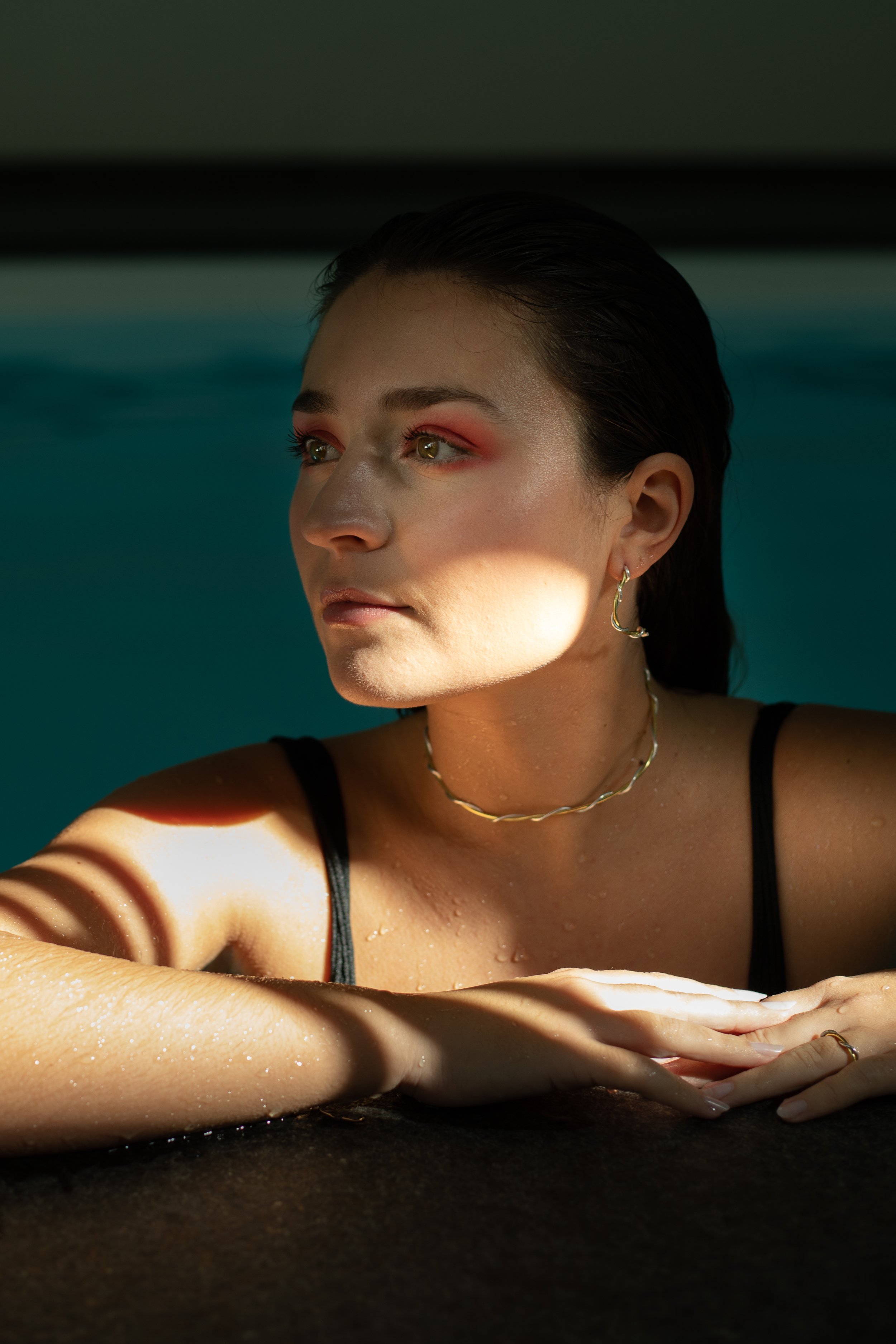 Une femme allongée dans une piscine, avec un éclairage subtil et des reflets, portant des boucles d'oreilles en argent, un collier, et un maillot noir.