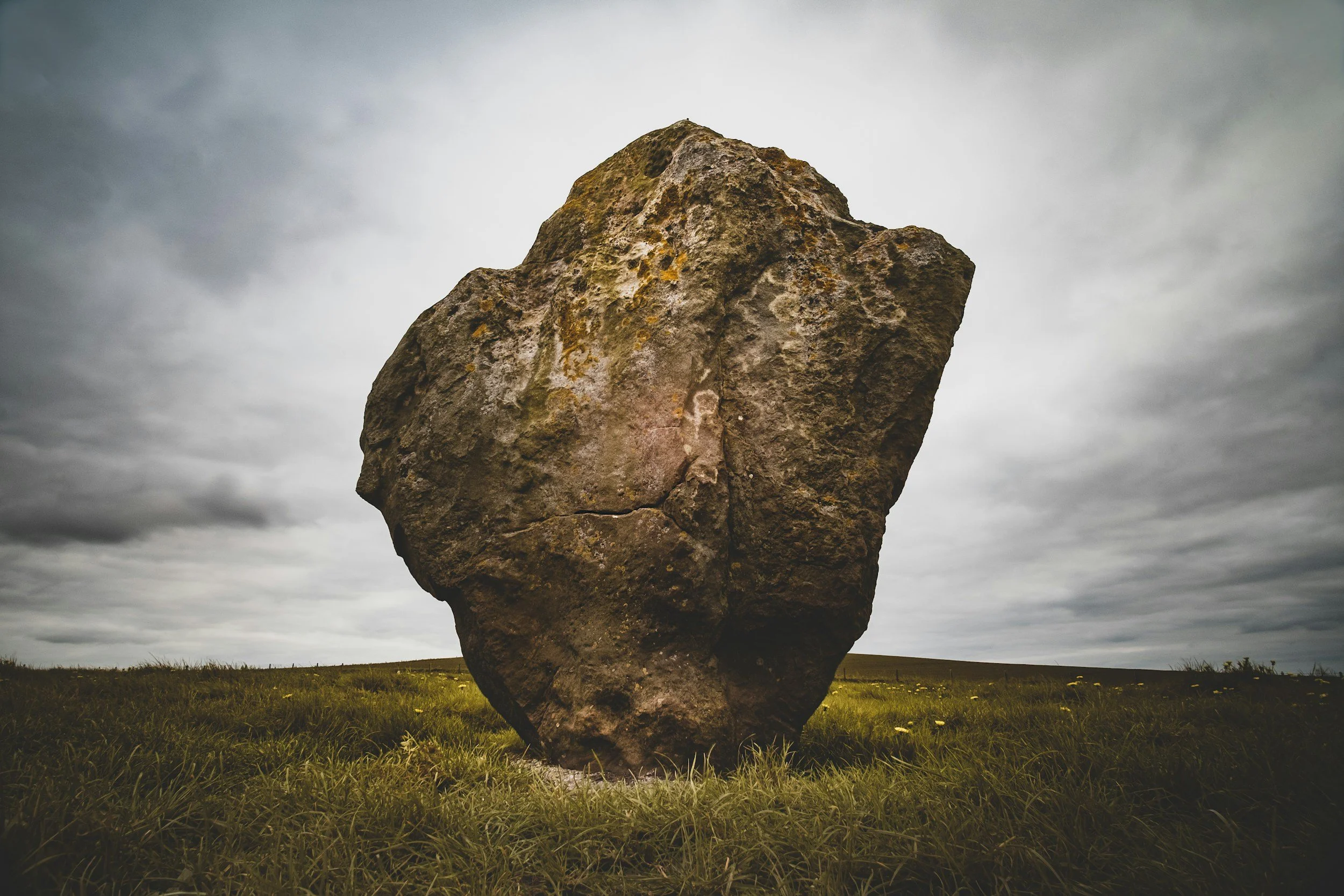 Large upright brown rock with a rough surface in a grassy field under a cloudy sky.
