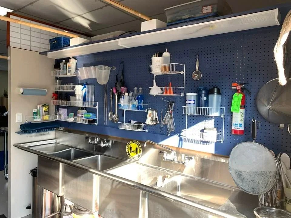 Commercial kitchen sink area with stainless steel double basin, mounted on a wall with dark blue pegboard. Wall-mounted shelves and racks hold cleaning supplies, utensils, and containers. A fire extinguisher is attached nearby, and sunlight shines on the workspace.