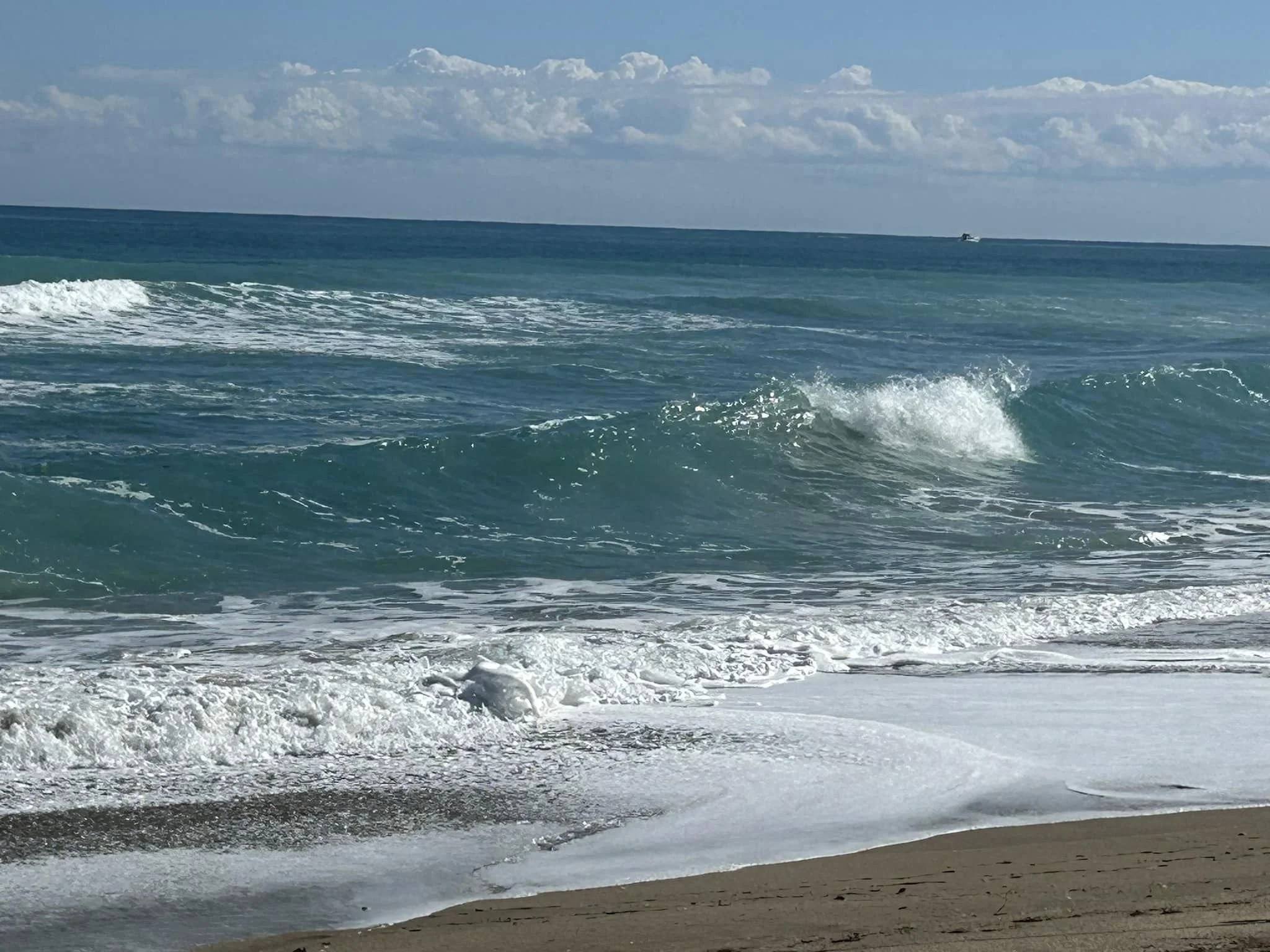 Ocean waves crashing on sandy beach under cloudy blue sky.