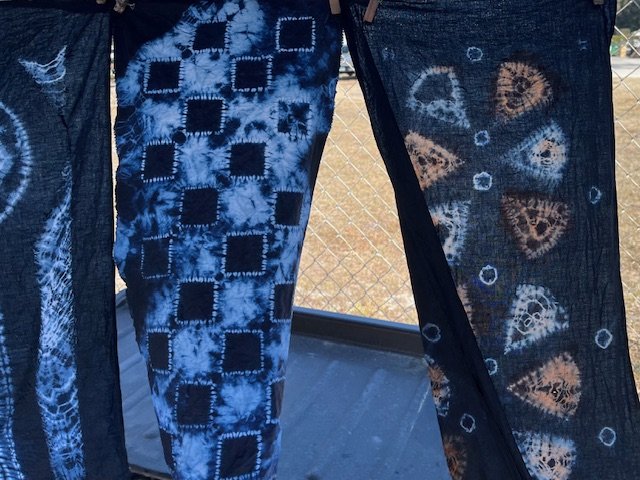 “Indigo tie-dyed fabric panels hanging outdoors with geometric square and circular patterns in blue and white.”