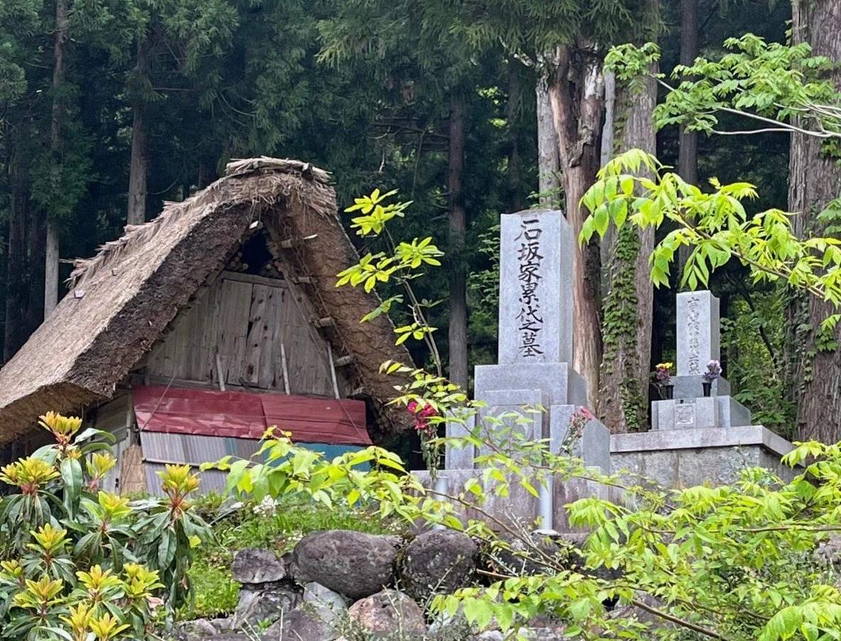A small Japanese shrine , surrounded by a thatched roof,  green foliage and trees, with two stone graves nearby.