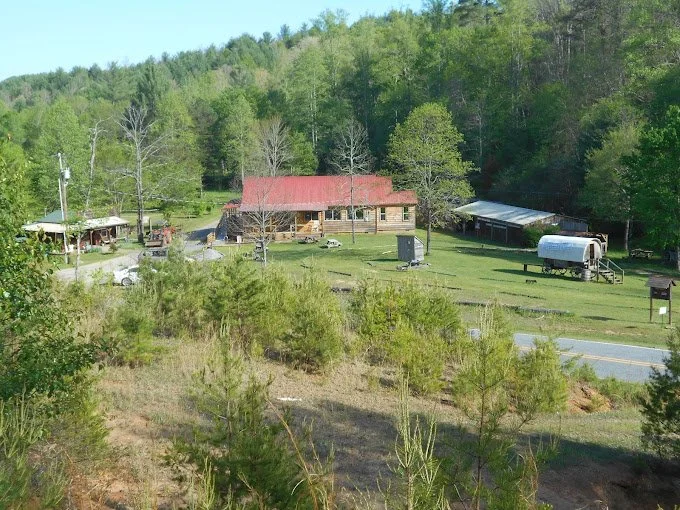 A rural scene featuring a house with a red roof, a forested hillside in the background, and a grassy area with trailers and small structures, near a road.