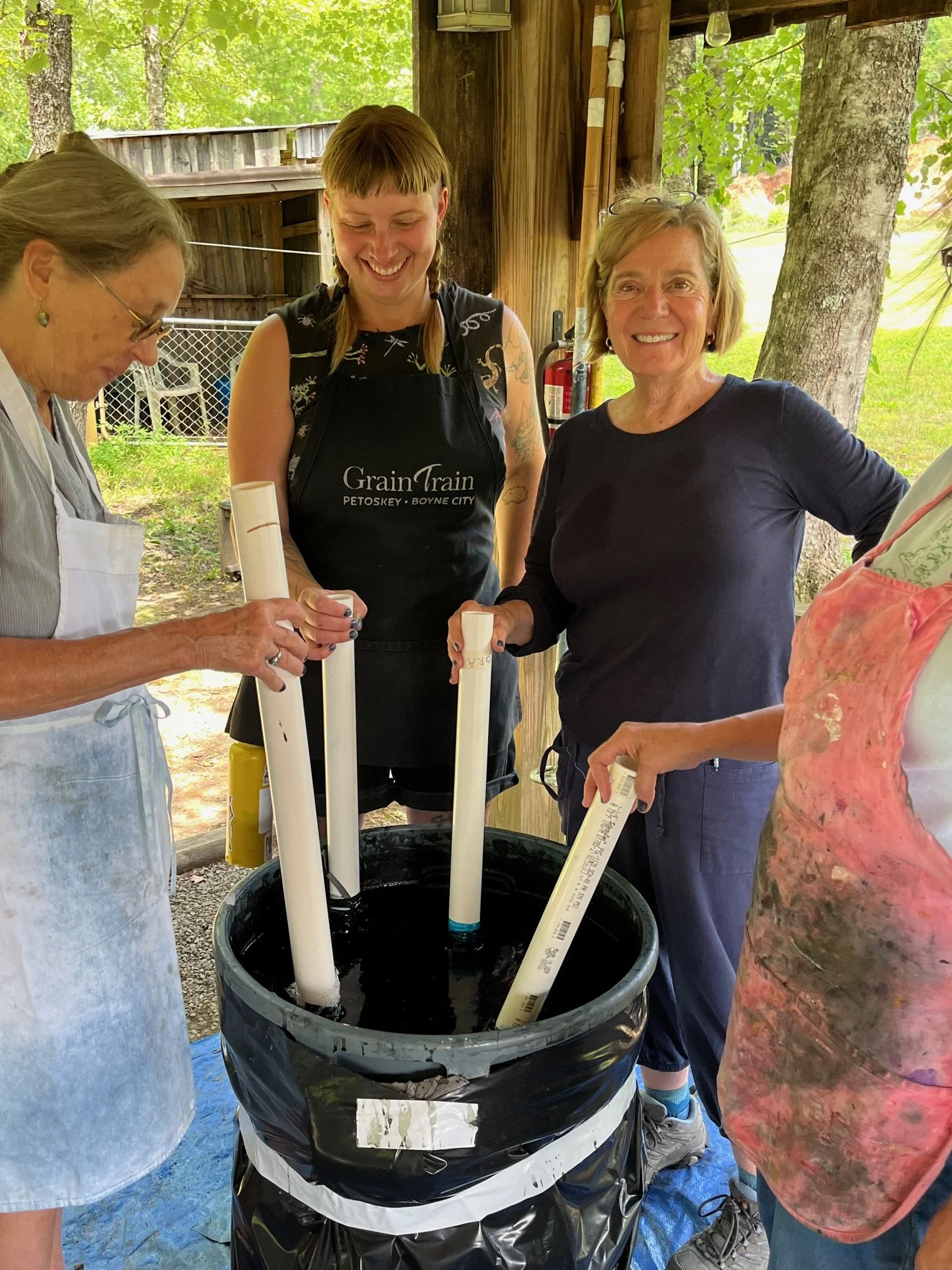 Group of women planting tubes into a black barrel in an outdoor wooden shelter surrounded by trees.