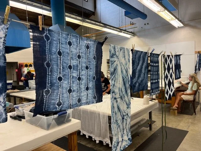 Various pieces of indigo-dyed fabric with traditional shibori patterns hanging on a line in a workshop setting.