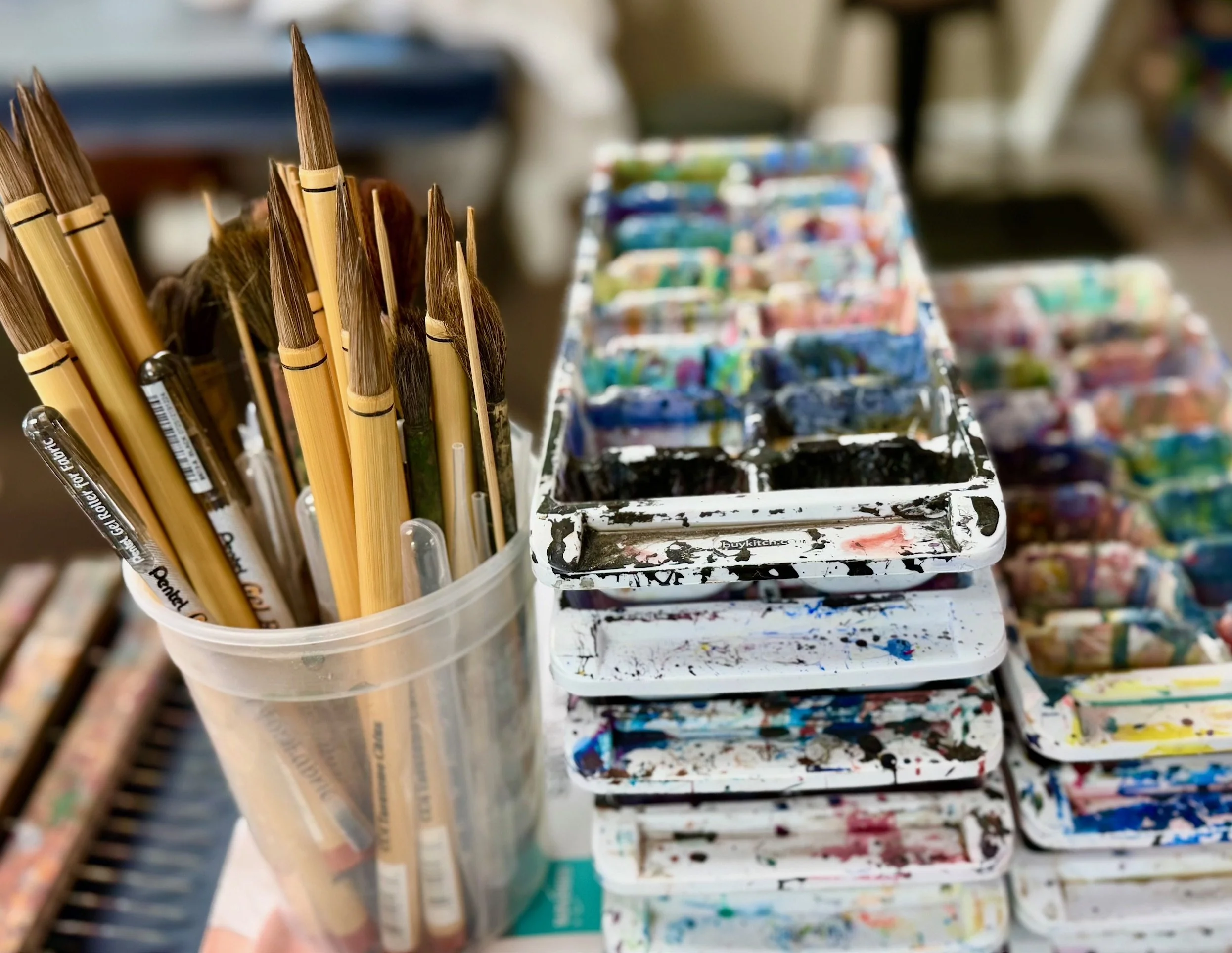 Paintbrushes in a container beside paint-stained trays in a working fiber art studio.