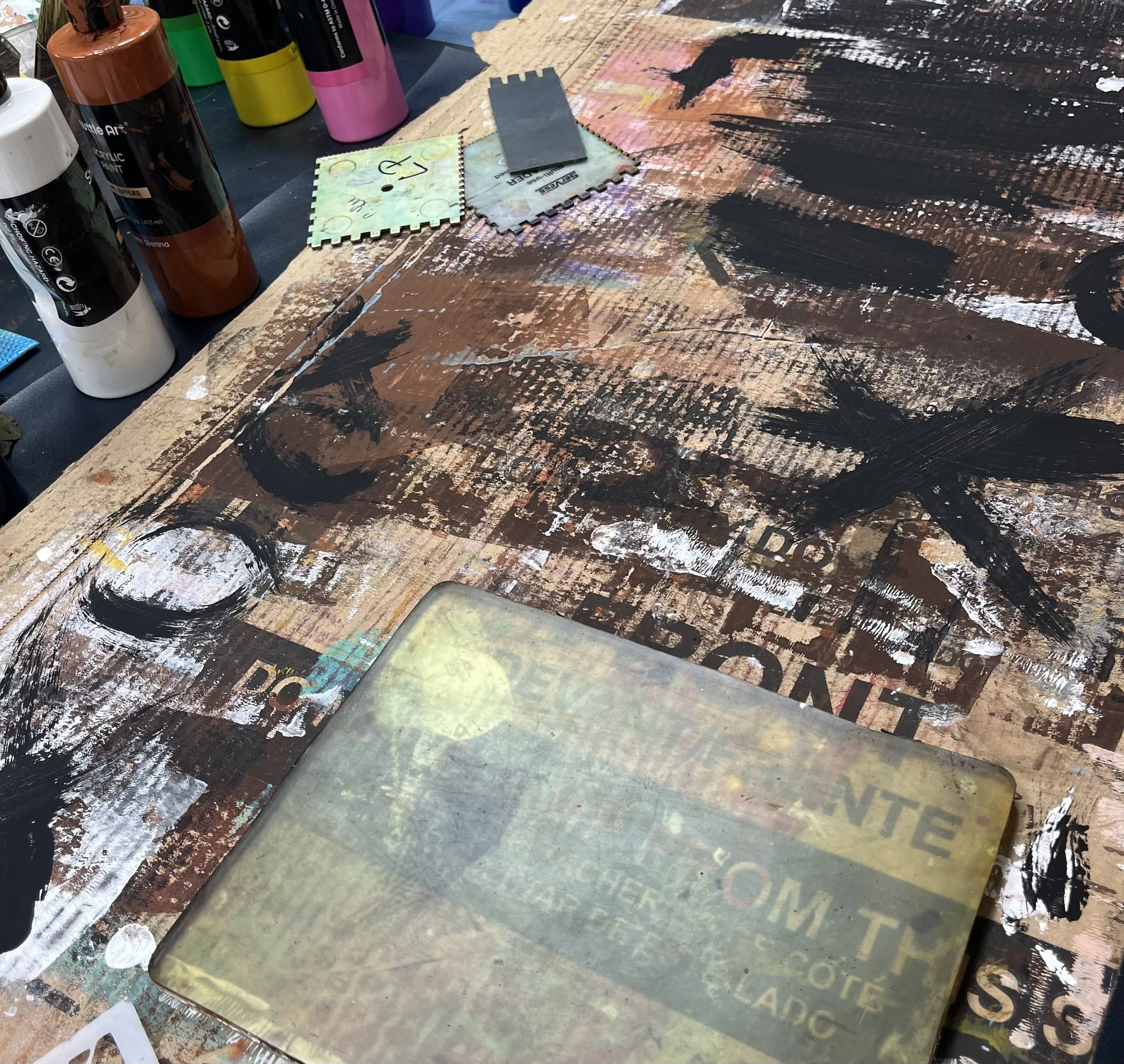 A cluttered art workspace with spray paint bottles, paint samples, and a large canvas covered in black, white, and brown abstract brush strokes.