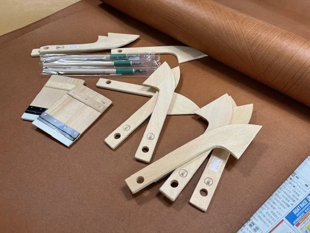 Traditional katazome tools including hand-carved wooden spatulas and fine brushes arranged on a brown work surface, used for Japanese stencil dyeing.