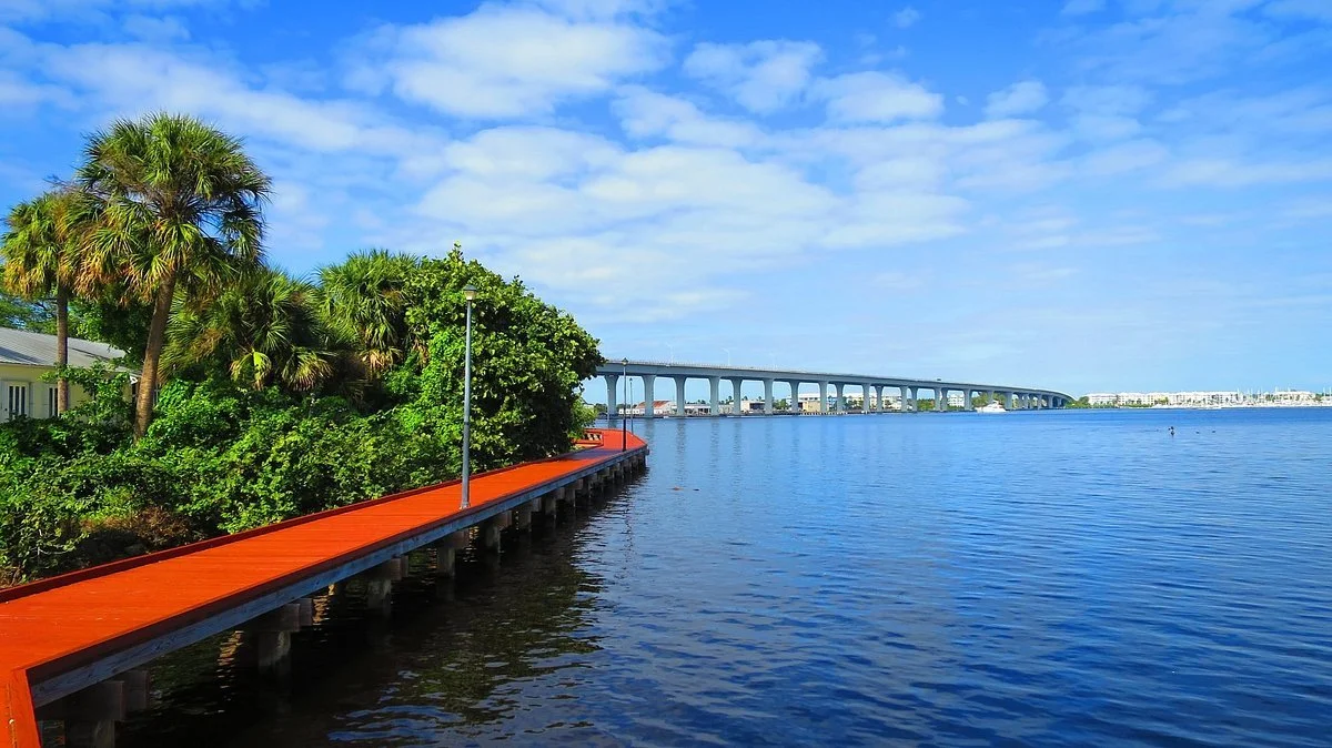 A waterfront scene with a wooden pier, lush green palm trees and bushes, and a bridge across a calm body of water under a partly cloudy blue sky.
