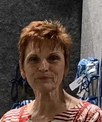 A woman with short reddish-brown hair wearing a red and white batik shirt, standing in front of a dark background with blue and white patterned fabric in the background.