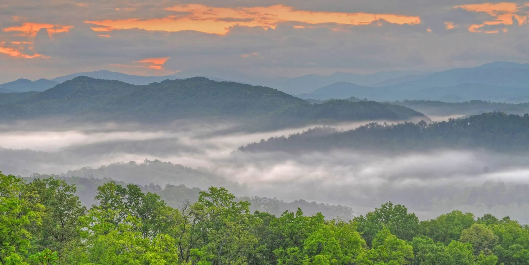 A landscape of lush green trees in the foreground, layers of misty mountains in the background, and an orange and pink sunset sky with dark clouds.