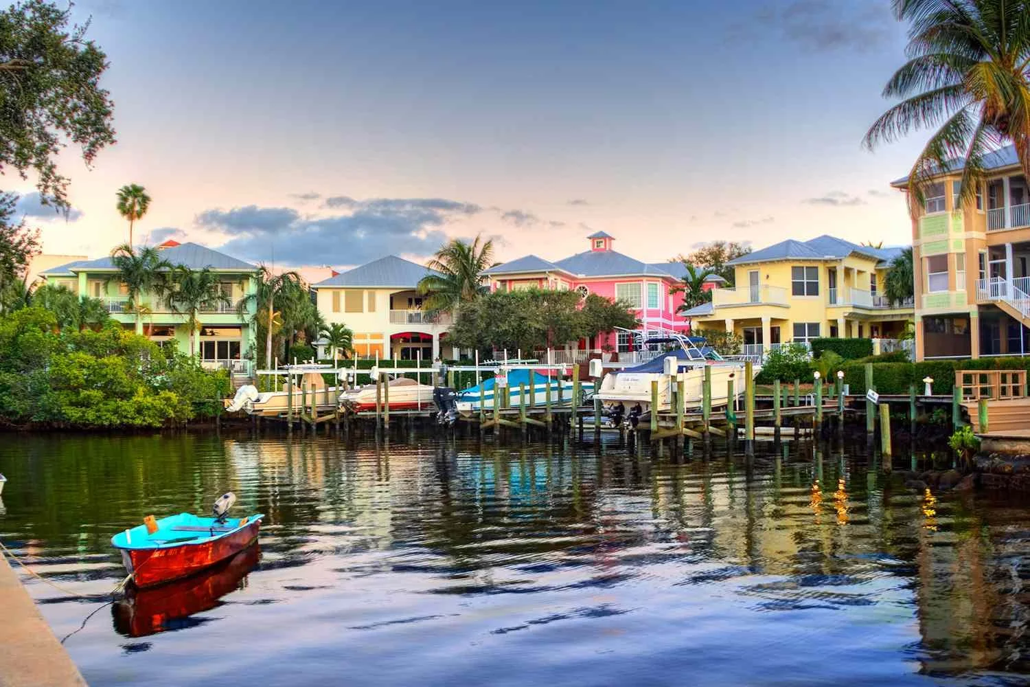 Colorful houses along a waterway with a small boat in the foreground, palm trees, and a dock during sunset.