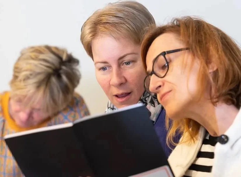 Three women looking at a book or menu together, with two in focus and one blurred in the background.