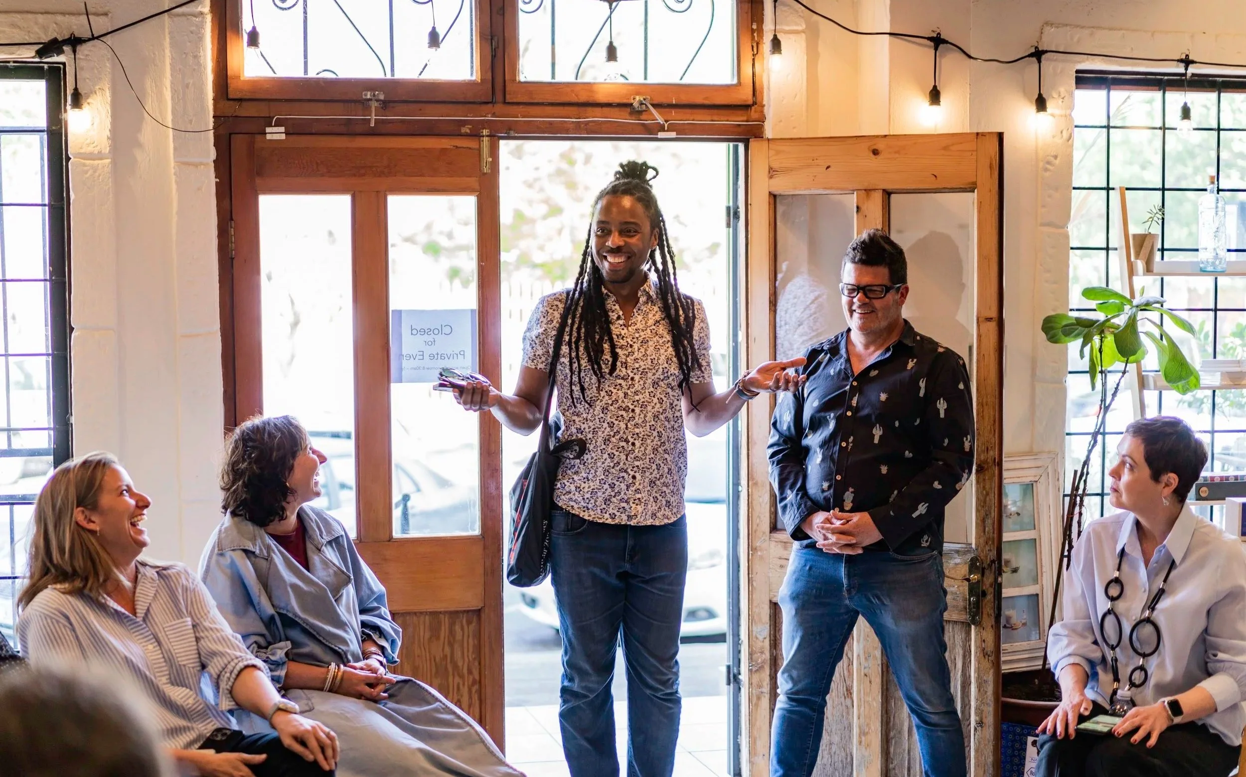 A diverse group of people are gathered inside a cafe or similar space, engaging in conversation. One person is standing and speaking, smiling, while others seated around are listening and laughing.