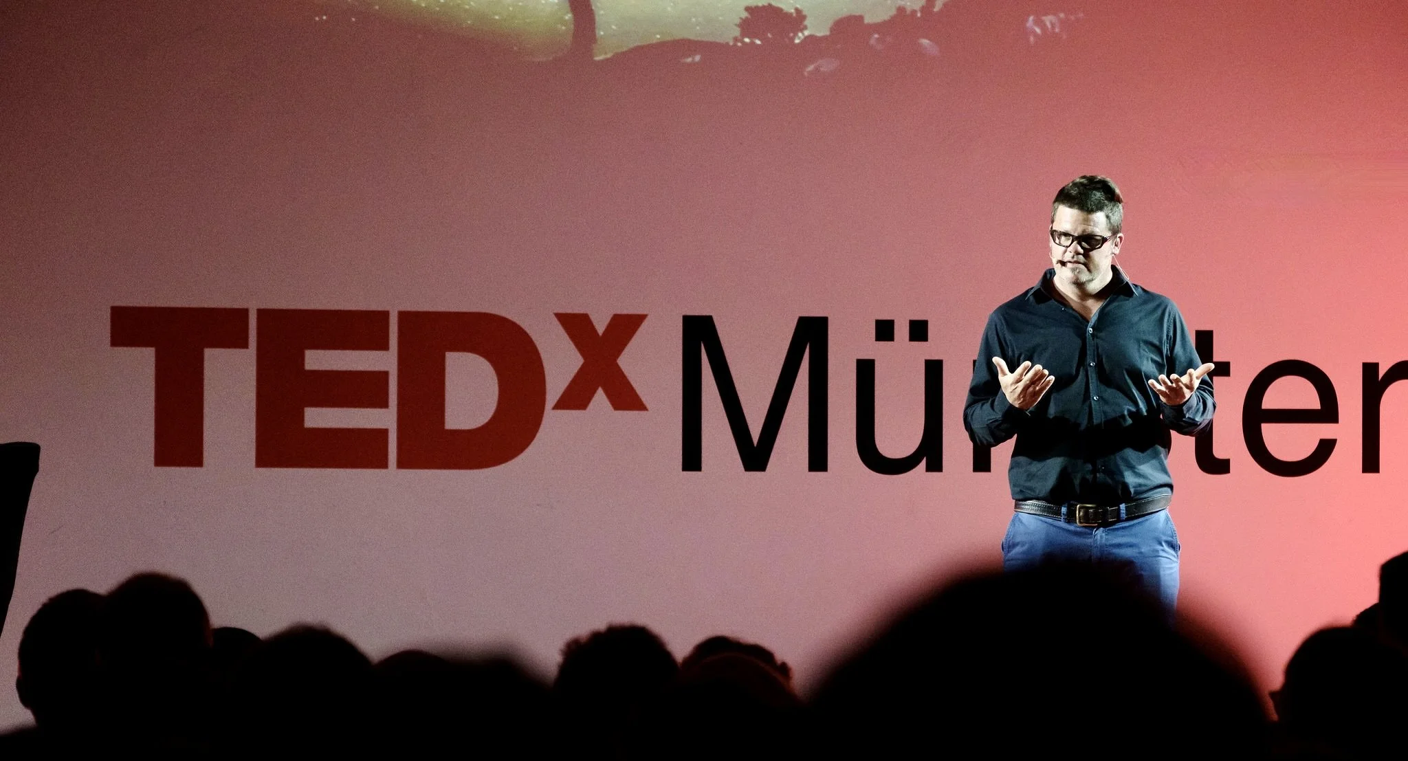 A man wearing glasses and a dark blue shirt giving a talk on stage at a TEDx event. The background has large red and black text reading 'TEDxMünster' with a pink gradient backdrop.