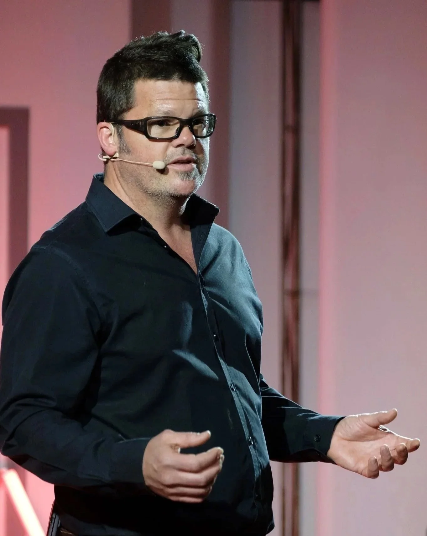 A man with dark hair, glasses, and a dark shirt speaking on stage with a headset microphone in front of a pink background.
