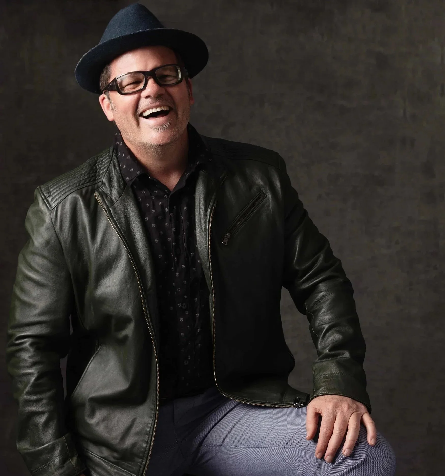 A smiling middle-aged man wearing a black fedora, glasses, a black leather jacket, a black shirt with small white dotted pattern, and gray pants, sitting against a dark background.
