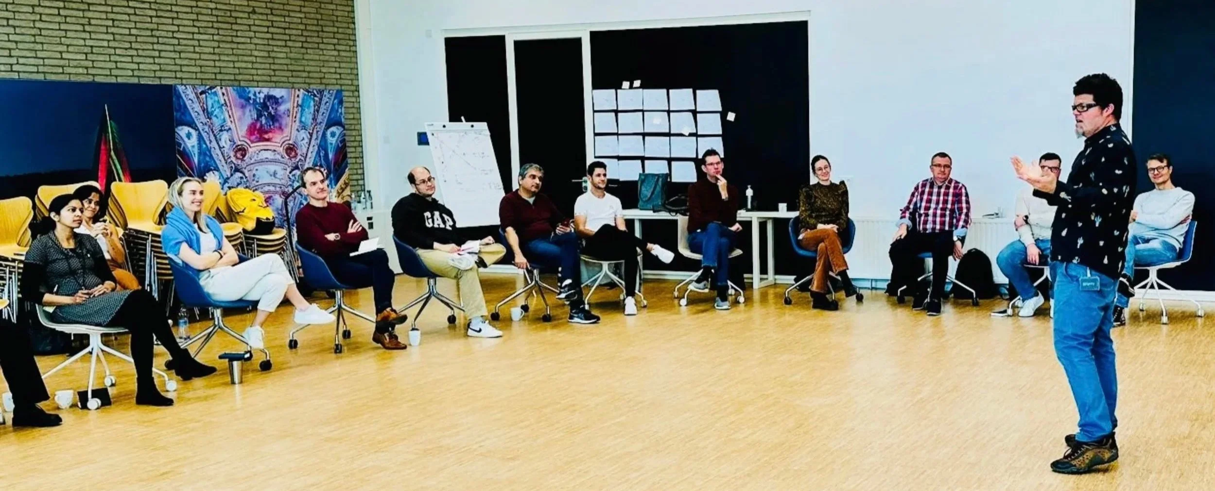 A man is giving a presentation to a group of 10 seated attendees in a bright room with wooden floors and black and white walls. The audience is sitting on chairs in a semi-circle, listening attentively.