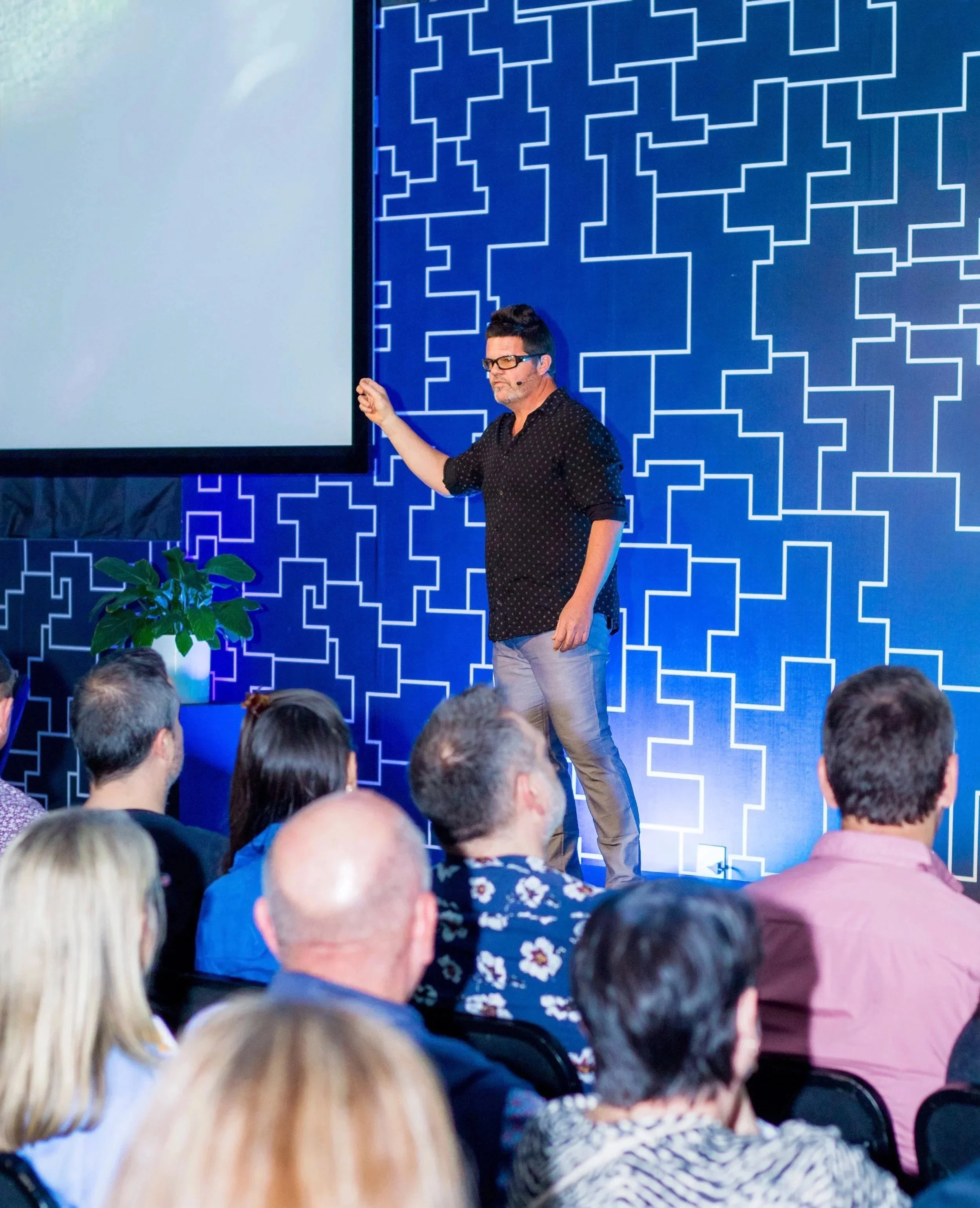A man standing on a stage giving a presentation in front of an audience. The background features a blue wall with a white geometric pattern, and there is a large screen to the left.
