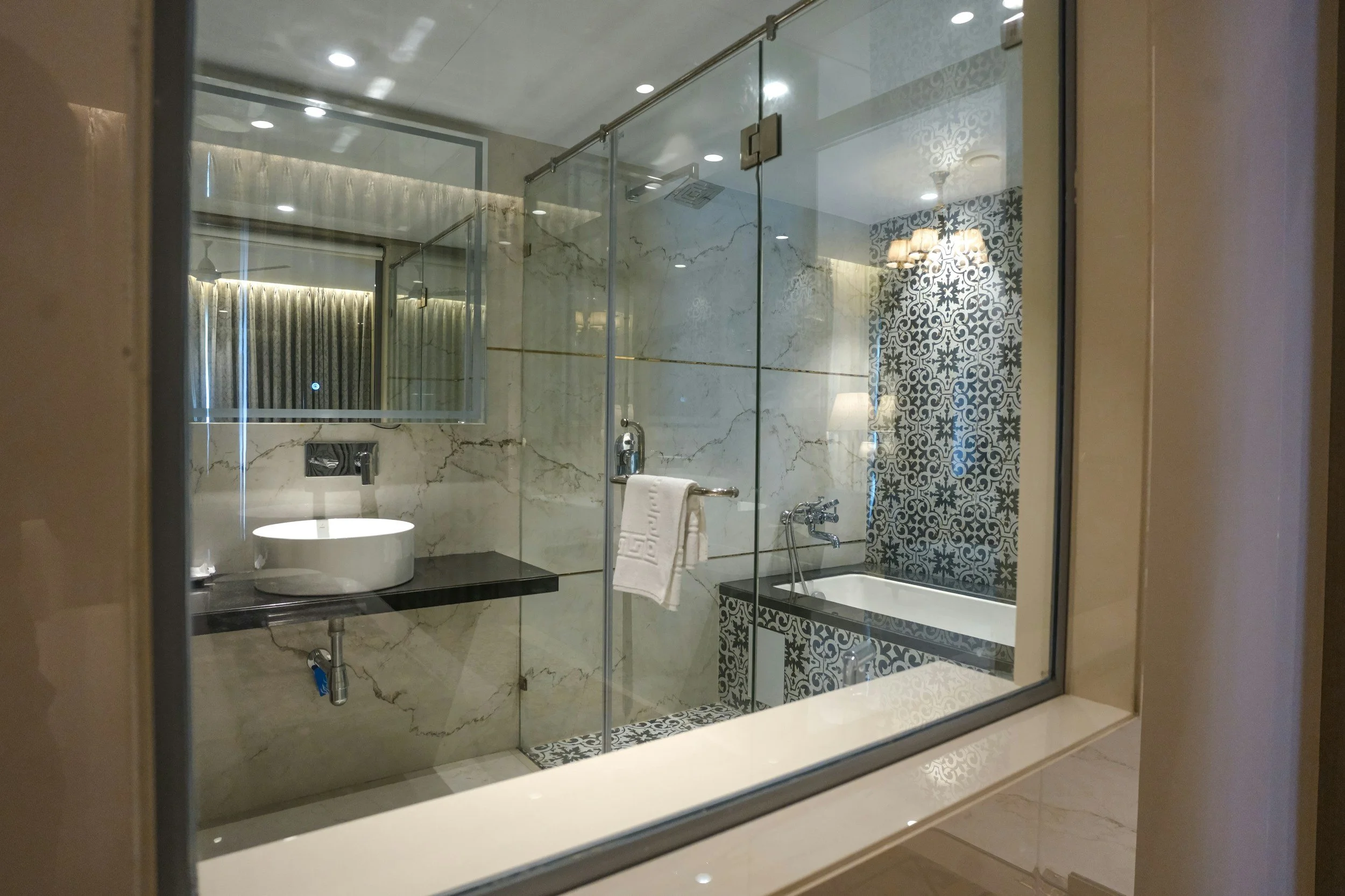 A modern bathroom with a glass shower enclosure, a black countertop with a round vessel sink, a wall-mounted faucet, a towel hanging from the glass shower door, a bathtub with chrome fixtures, and decorative tiles on the wall.
