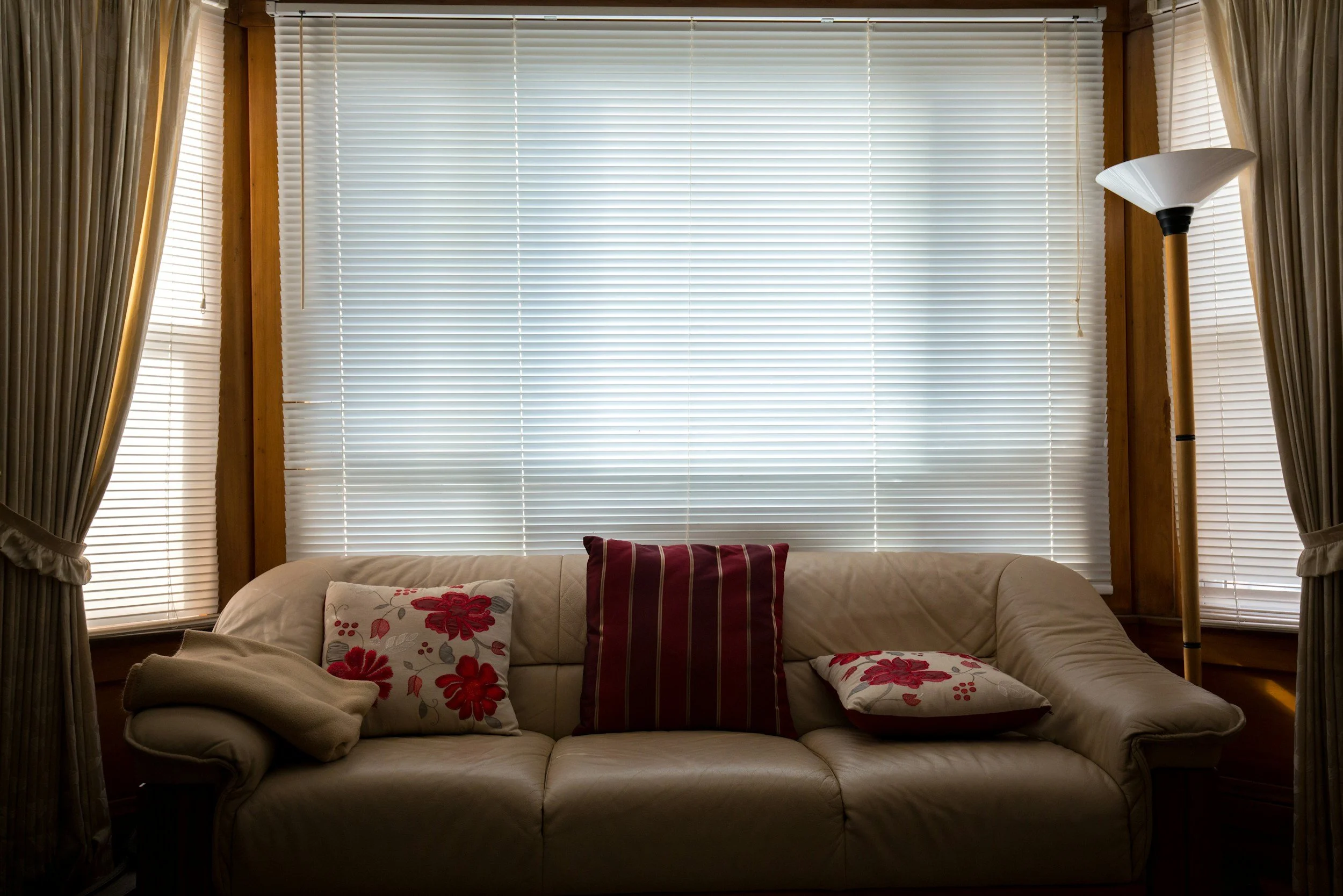 Living room with a beige sofa, three decorative pillows (two with floral designs, one striped), surrounded by large windows with white blinds and beige curtains, and a tall floor lamp with a white shade on the right side.