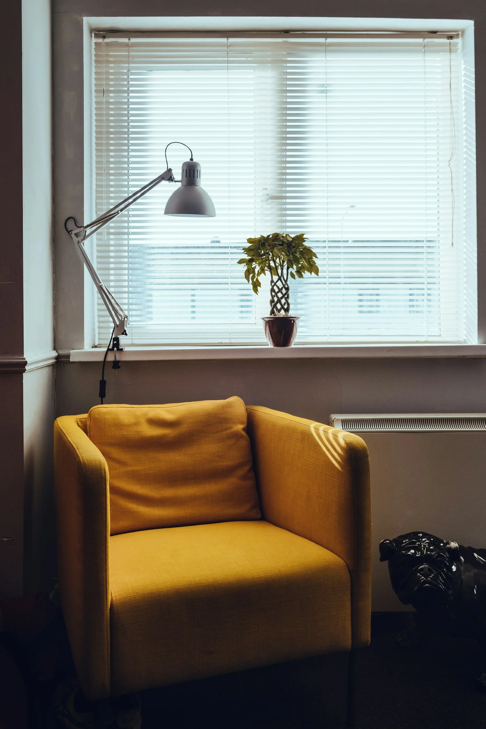 A cozy room corner with a yellow armchair, a window with horizontal blinds, a potted plant on the windowsill, a white adjustable desk lamp, and a black bulldog figurine on the floor.