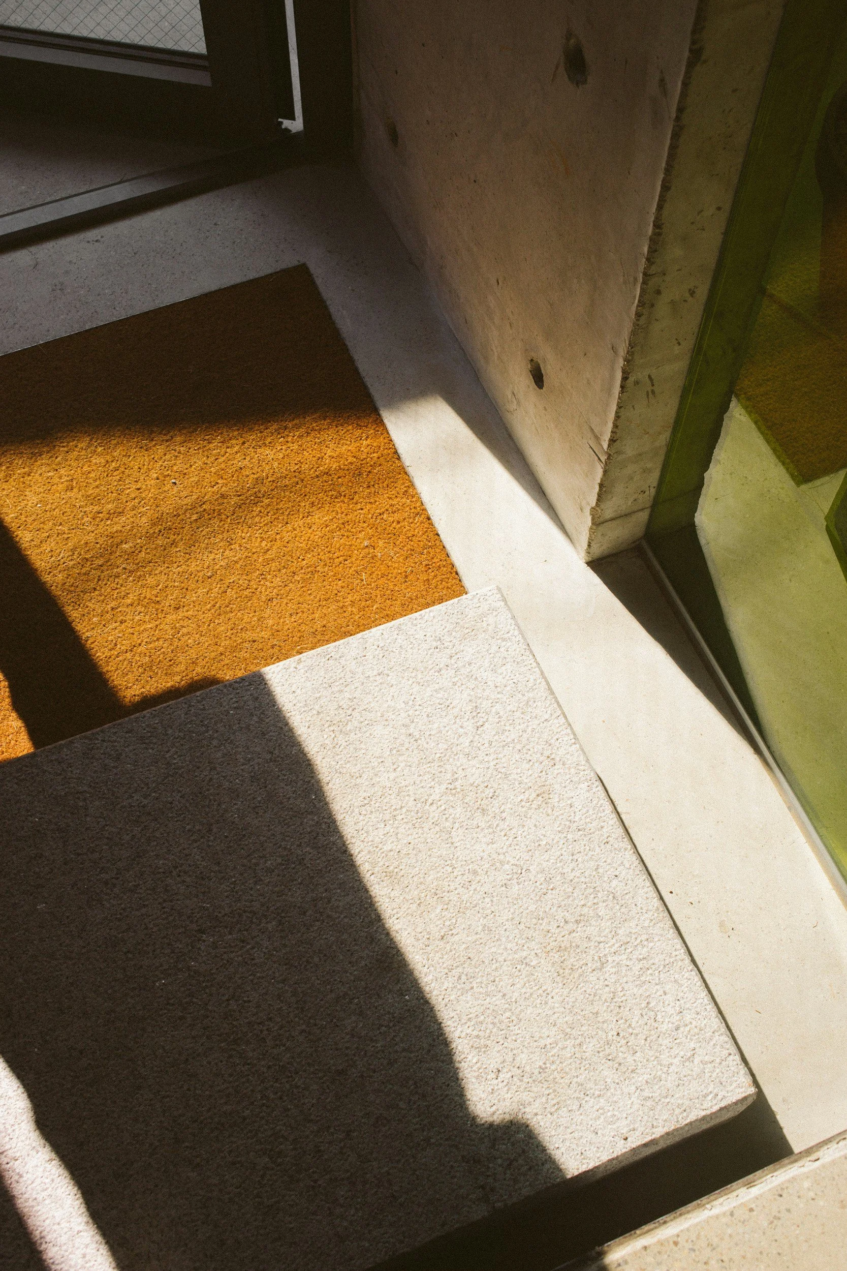 Close-up of entrance step with orange doormat, concrete flooring, and a door frame casting shadow.