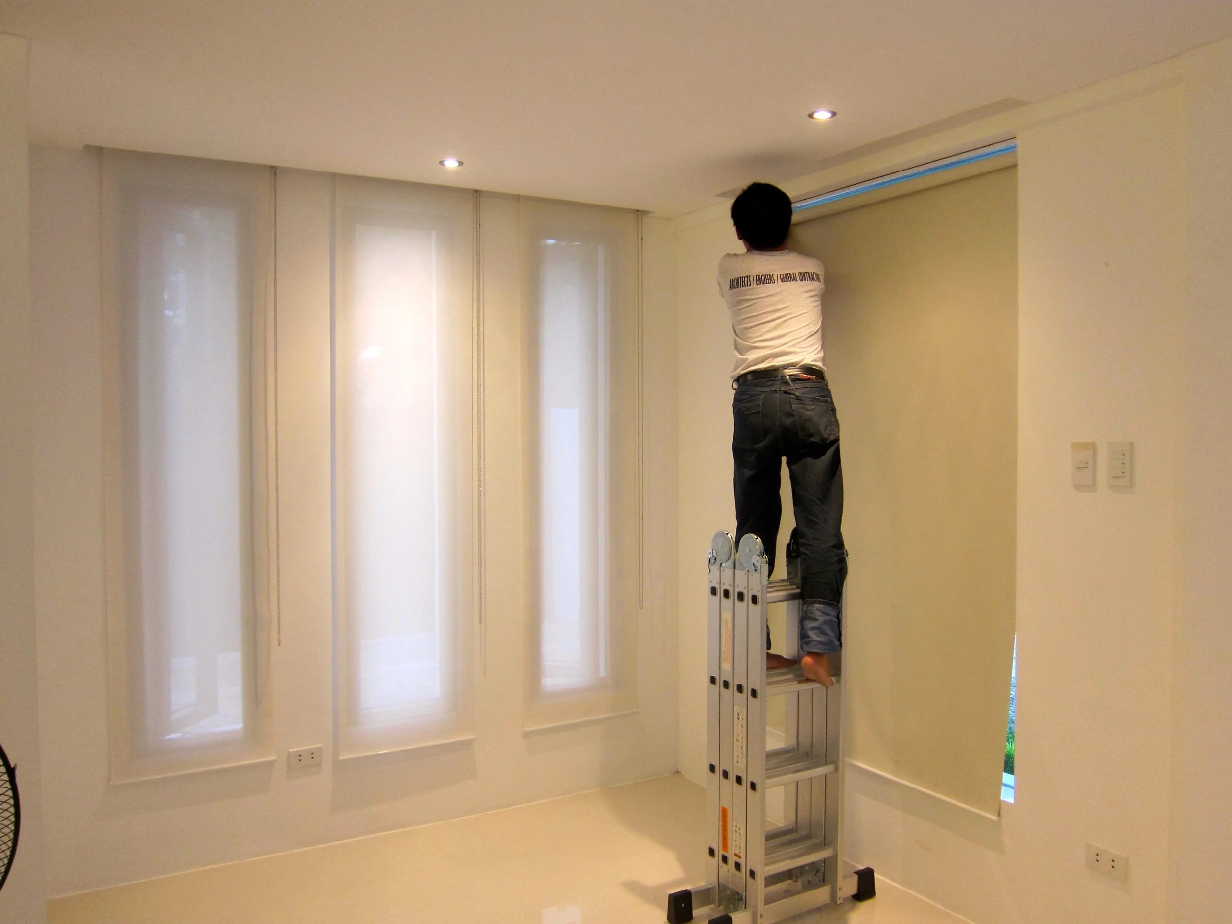 A person standing on a ladder working on a window or window shade in a room with white walls and beige curtains.