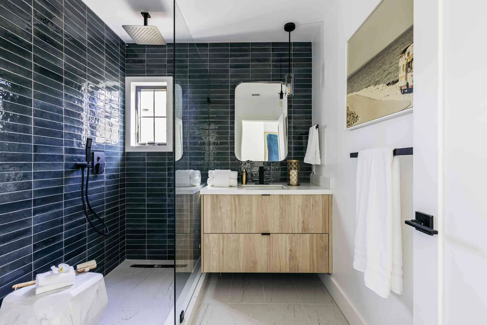 Modern bathroom with black tiled shower area, a small window, a wooden vanity with a mirror, white towels, and beach-themed artwork on the white wall.