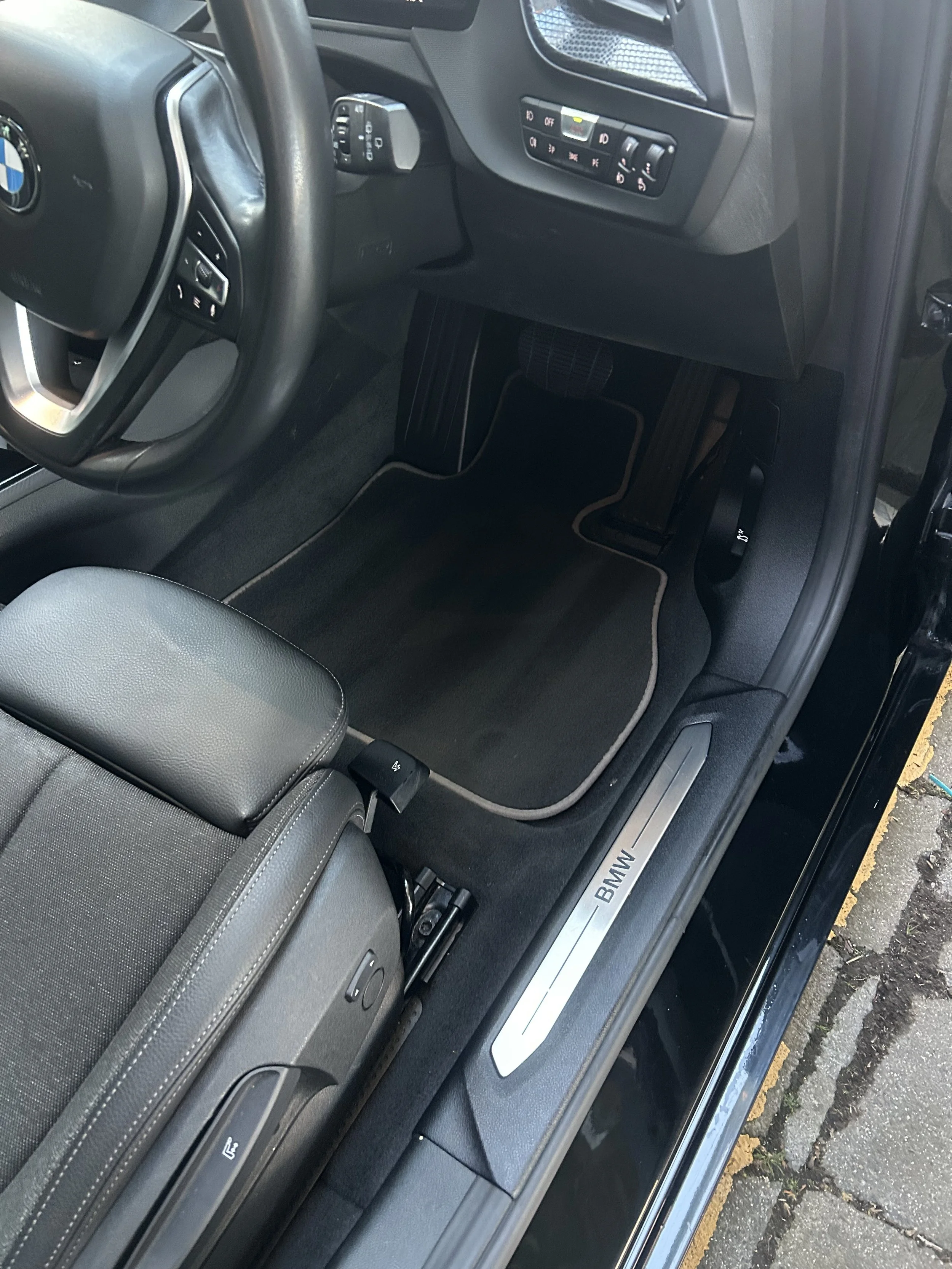 Interior of a BMW vehicle, showing the driver's side foot area, steering wheel, and door sill with BMW logo.