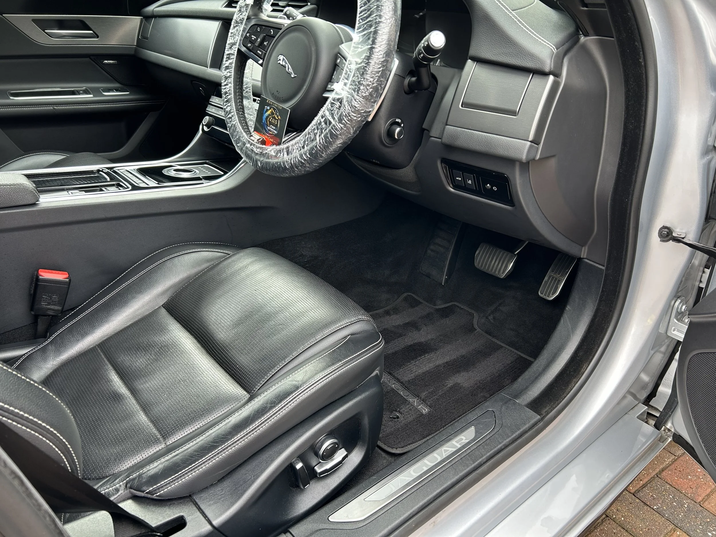 Interior of a Jaguar vehicle showing the driver's seat, steering wheel with a protective cover, dashboard, pedals, and floor mat with Jaguar logo.
