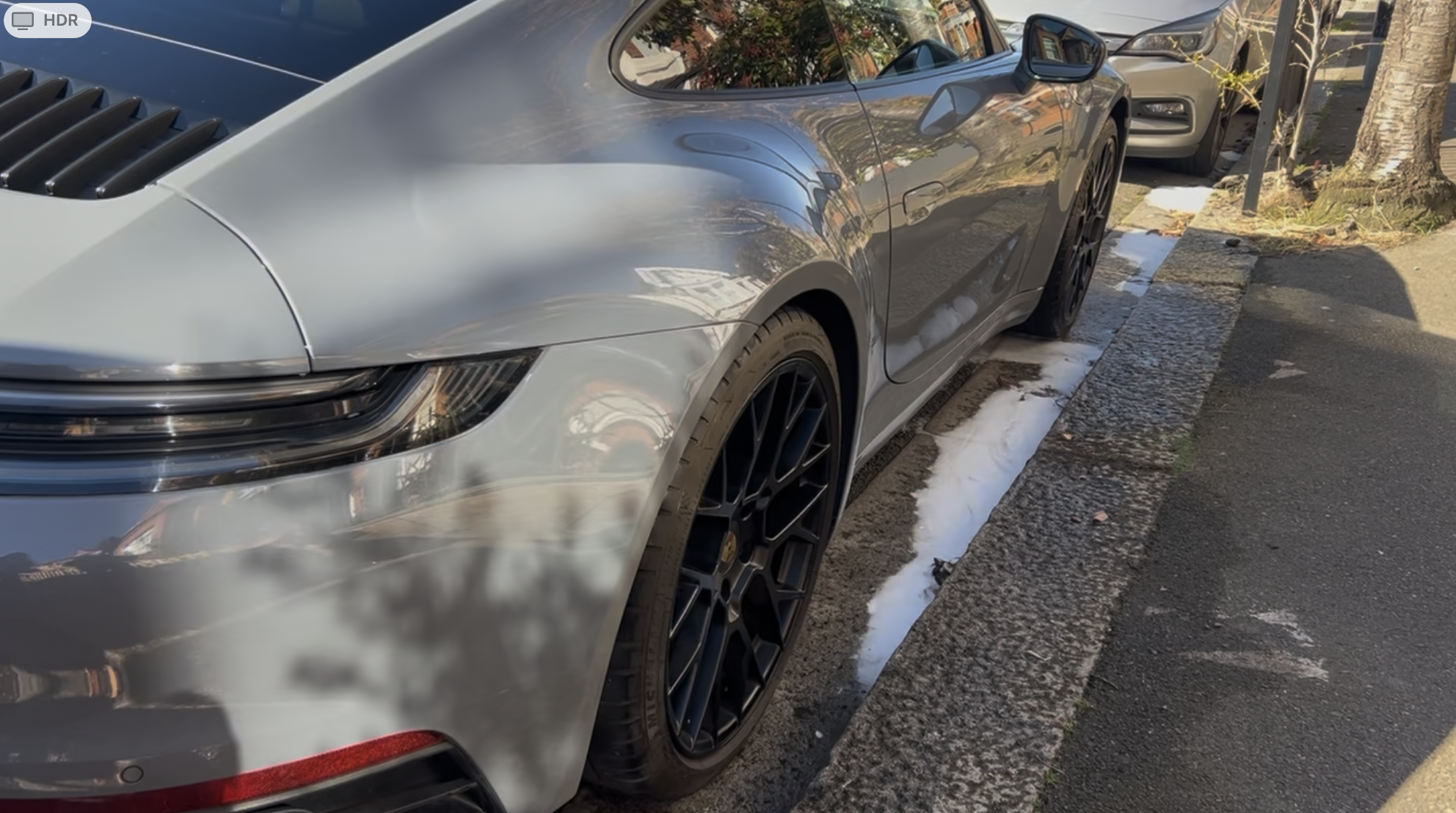 Front side of a silver Porsche sports car parked on the street with foam or soap on the ground beside it.