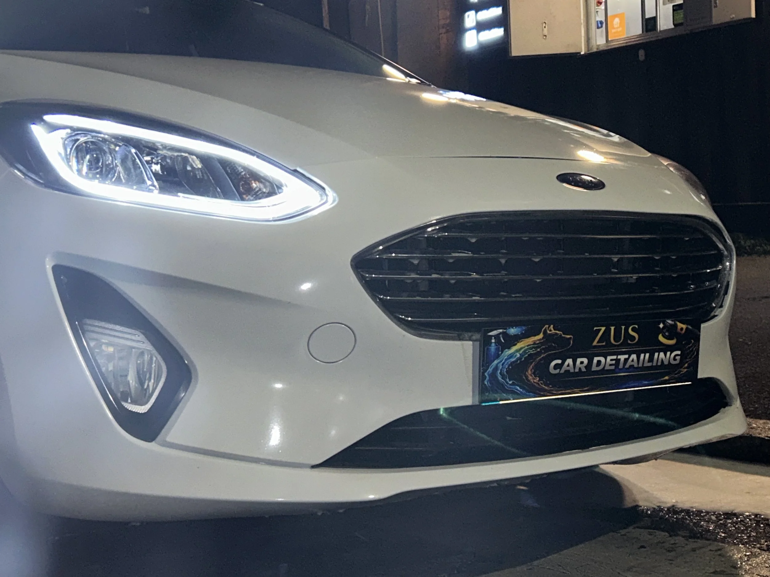 Front view of a white Ford vehicle with its headlights on, parked outdoors at night, with a car detailing service sign on the front bumper.