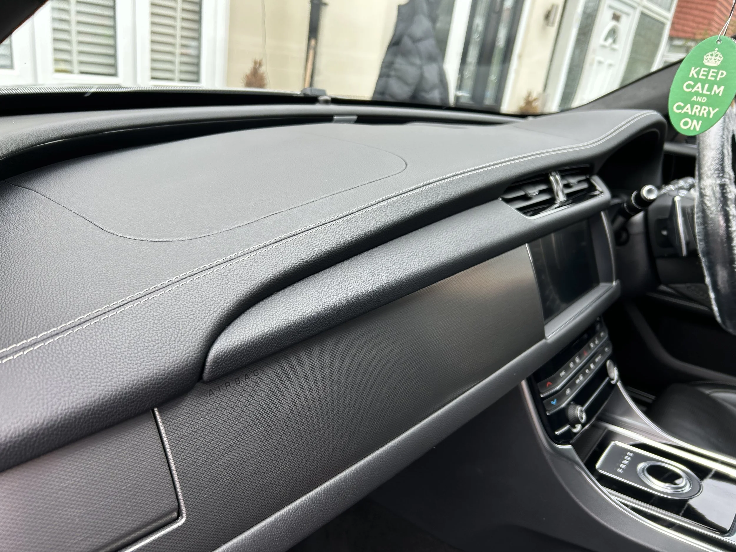 View of the dashboard of a modern car, showing the air vents, digital display, and climate control panel, with a green air freshener hanging from the rearview mirror.